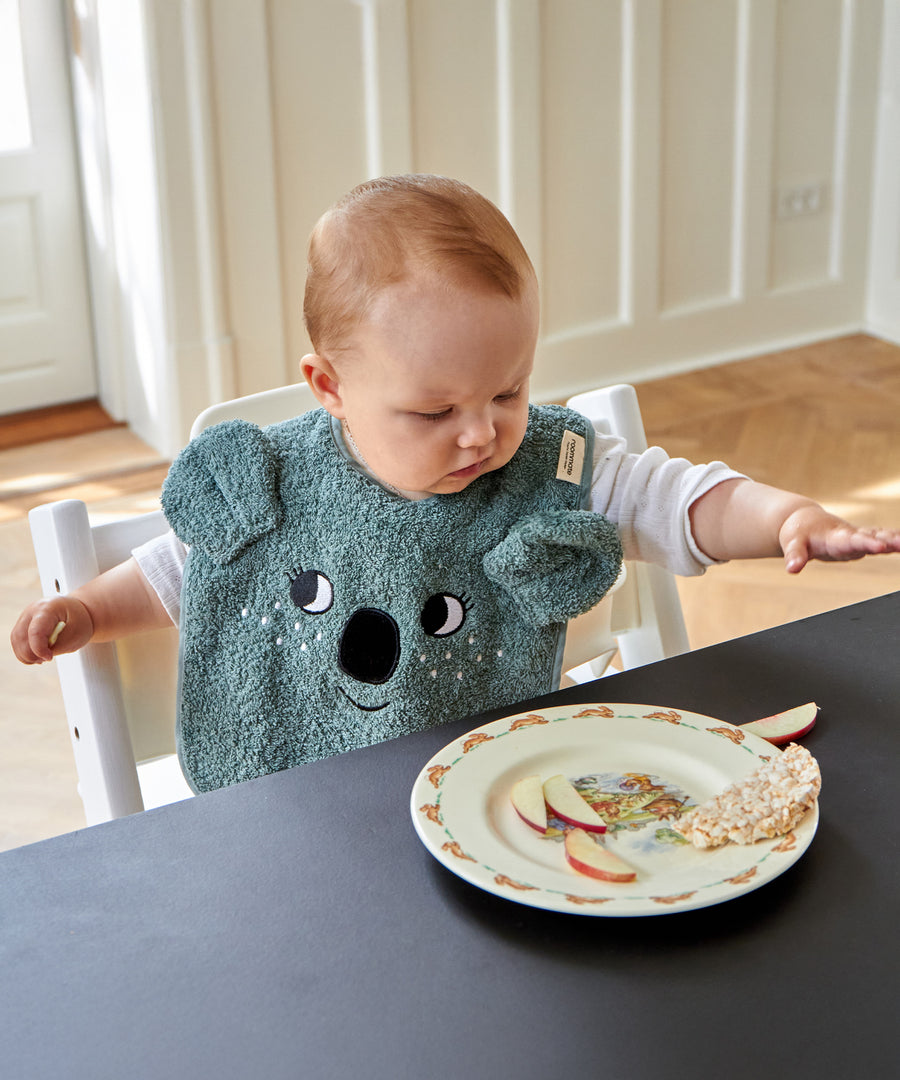 A child sat at a table whilst eating, wearing the Roommate Organic Cotton Baby Bib - Koala