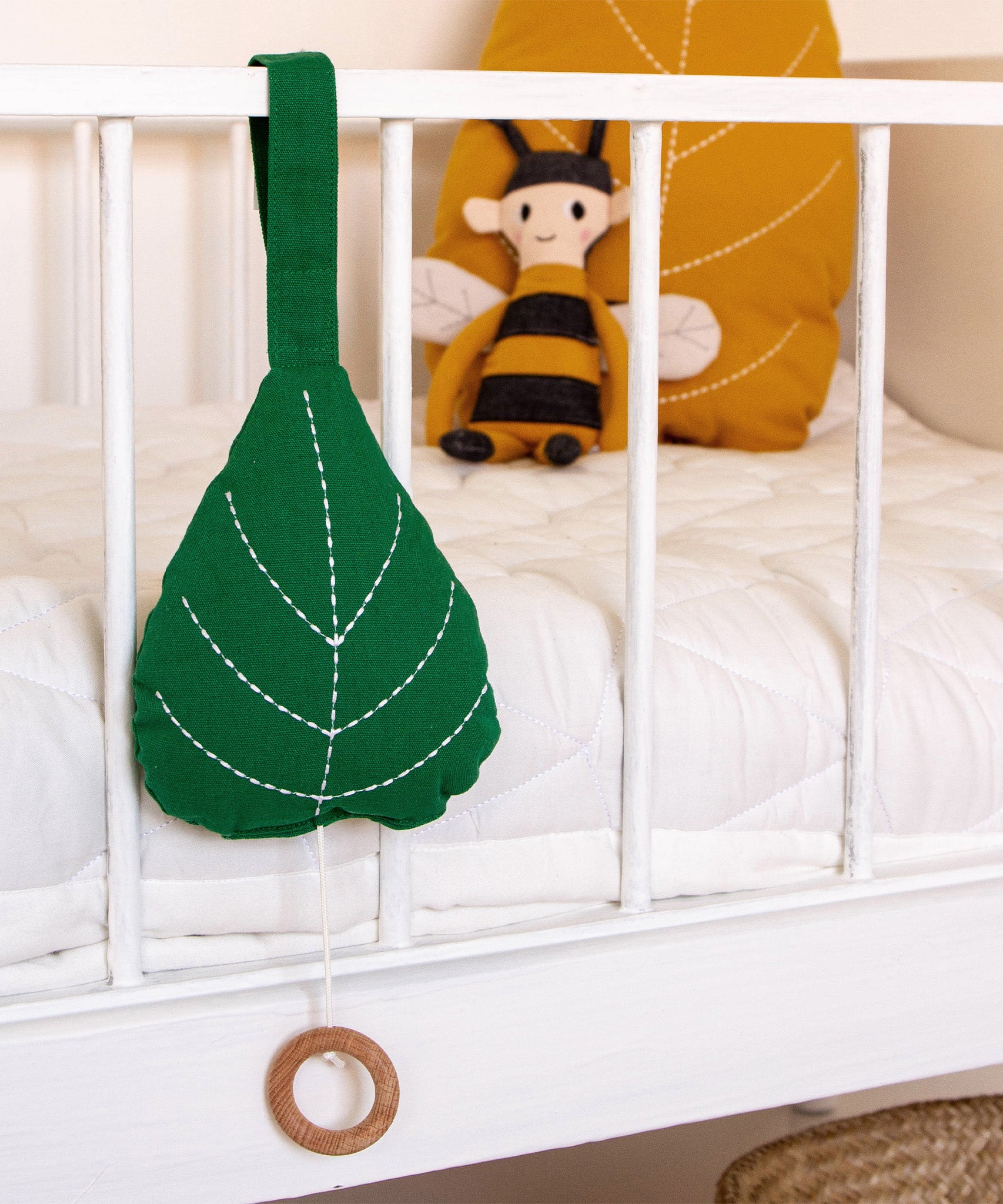 The Roommate Leaf Lullaby Music Mobile is attached to a white crib, and in the background is  Roommate yellow leaf cushion and a Roommate Bee Rag Doll