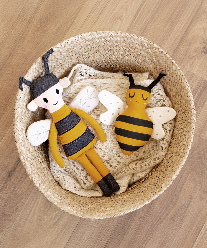 The Roommate Organic Cotton Bee Rattle in a basket with another Roommate bee toy, on a wooden floor background