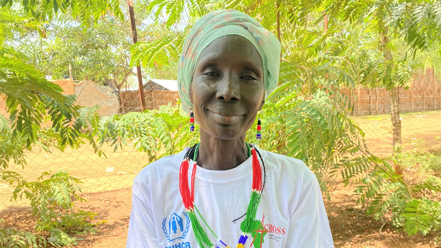 Hana, a smiling refugee artisan, stands outdoors wearing a white t-shirt, proud of her work in the #WithRefugees project