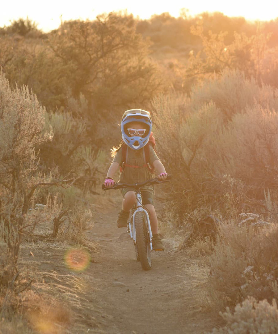 A child riding the Early Rider Seeker X16 16" Bike - Brushed Aluminium through shrubs and trees, on a dirt trail