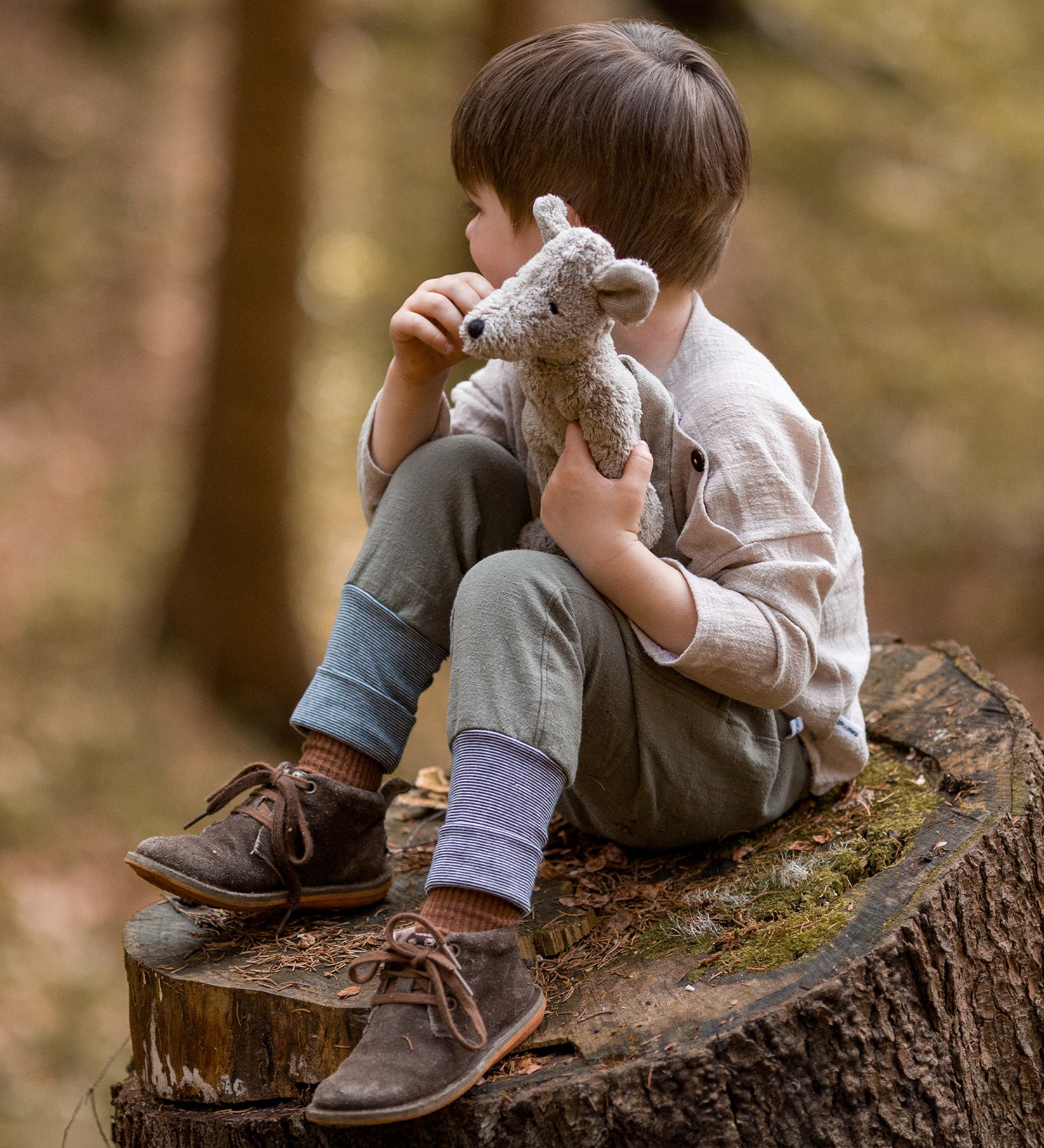 A child is sat on a large, cut tree stump, looking off into the distance, whilst holding the Senger Baby Mouse