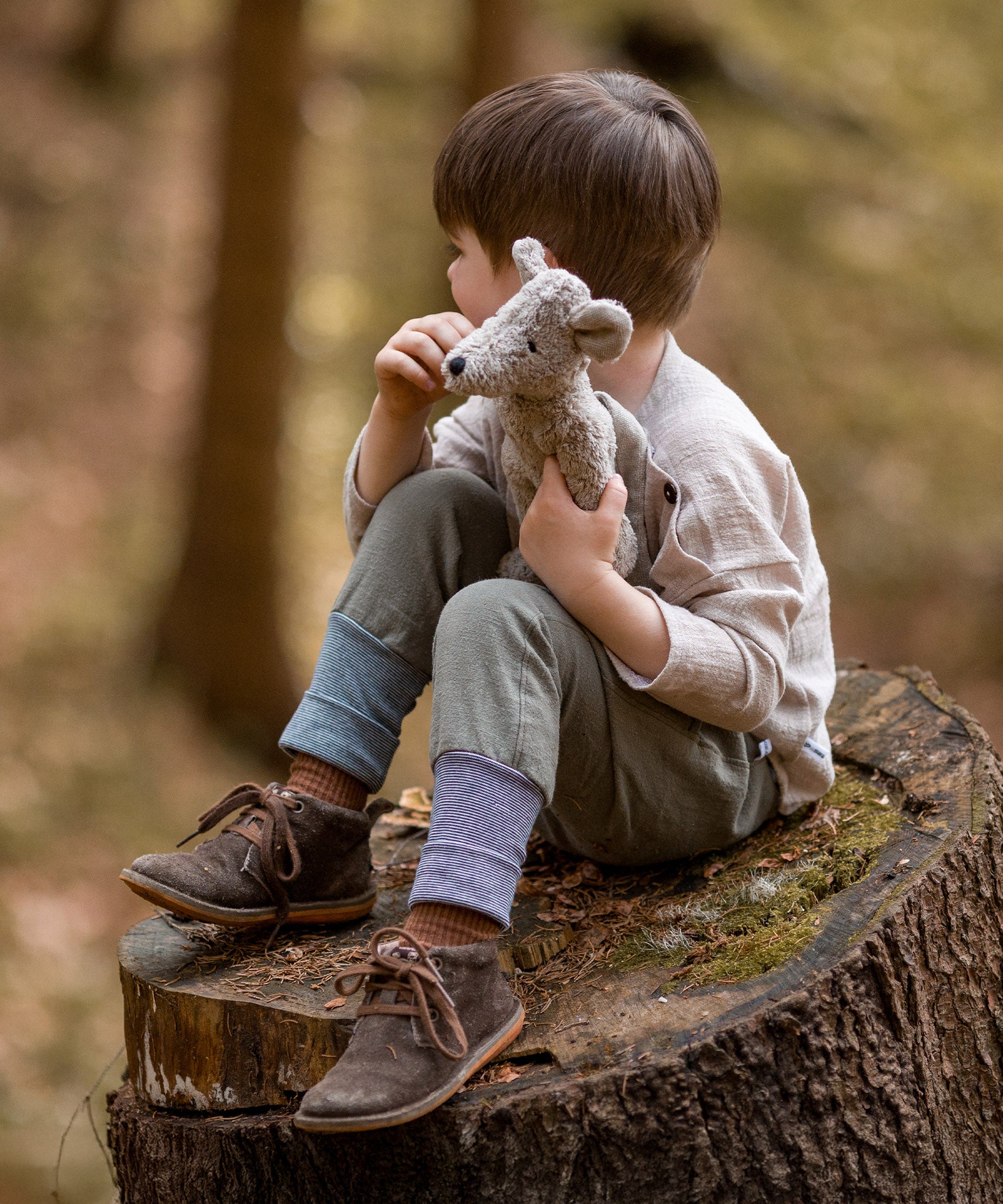 A child is sat on a large, cut tree stump, looking off into the distance, whilst holding the Senger Baby Mouse