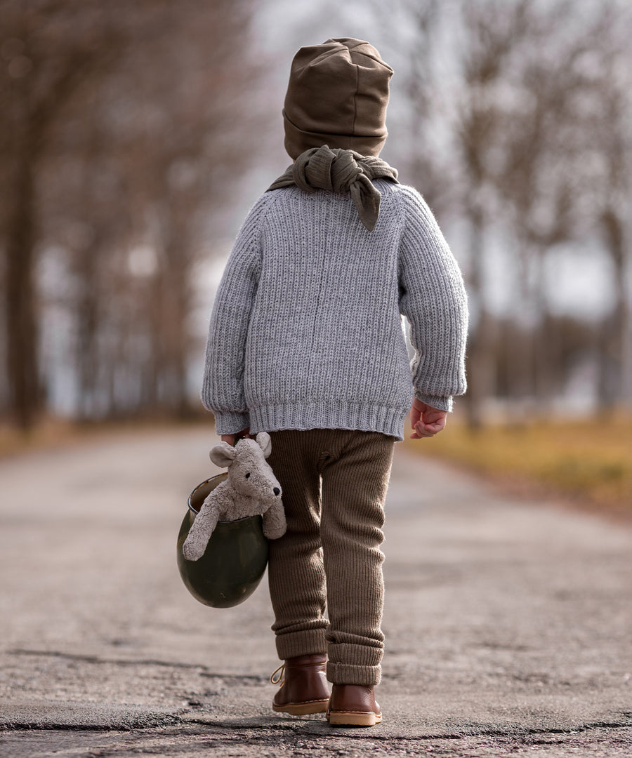 A child walking down a deserted road, whilst carrying the Senger Baby Mouse in a green jug with their left hand. The image shows the child from the back and the Senger Baby Mouse in the jug being held down by the child's left knee