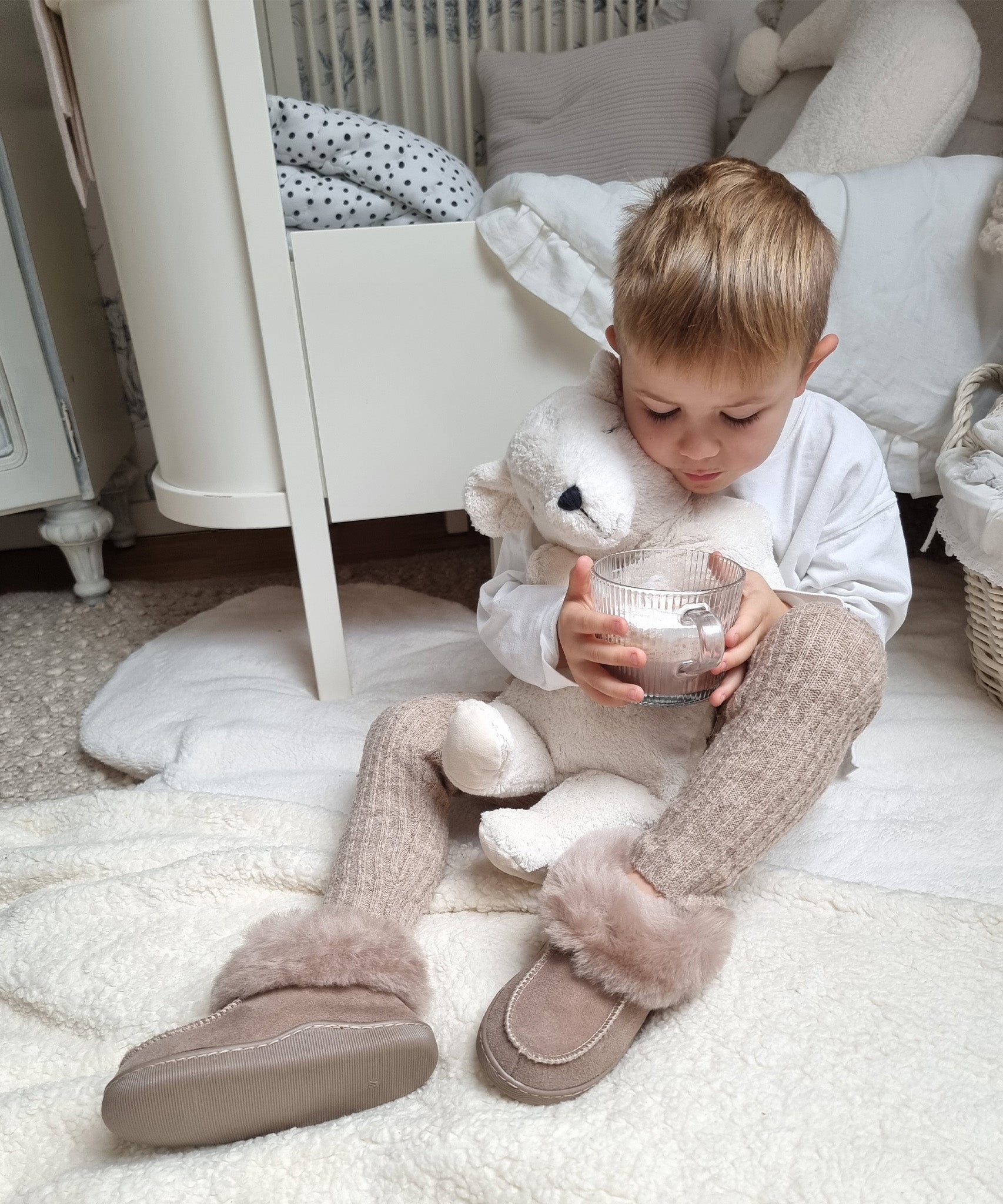 A child sat on a fluffy white rug in their room, holding the Senger Large Cuddly Polarbear in their arms and a cup of hot chocolate