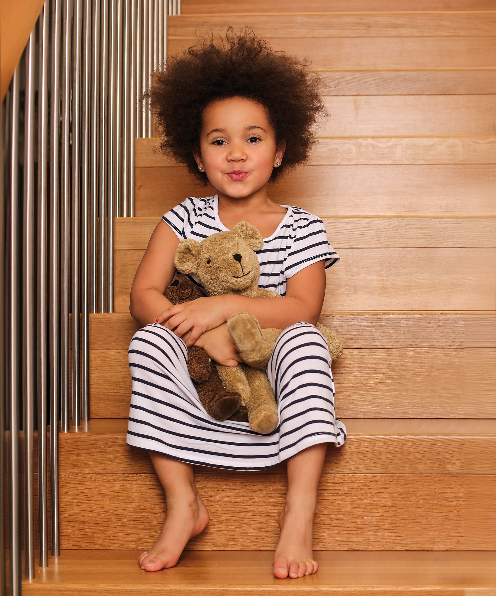 A child sat on wodden stairs, holding a Senger Large Floppy Beige Bear and small brown bear