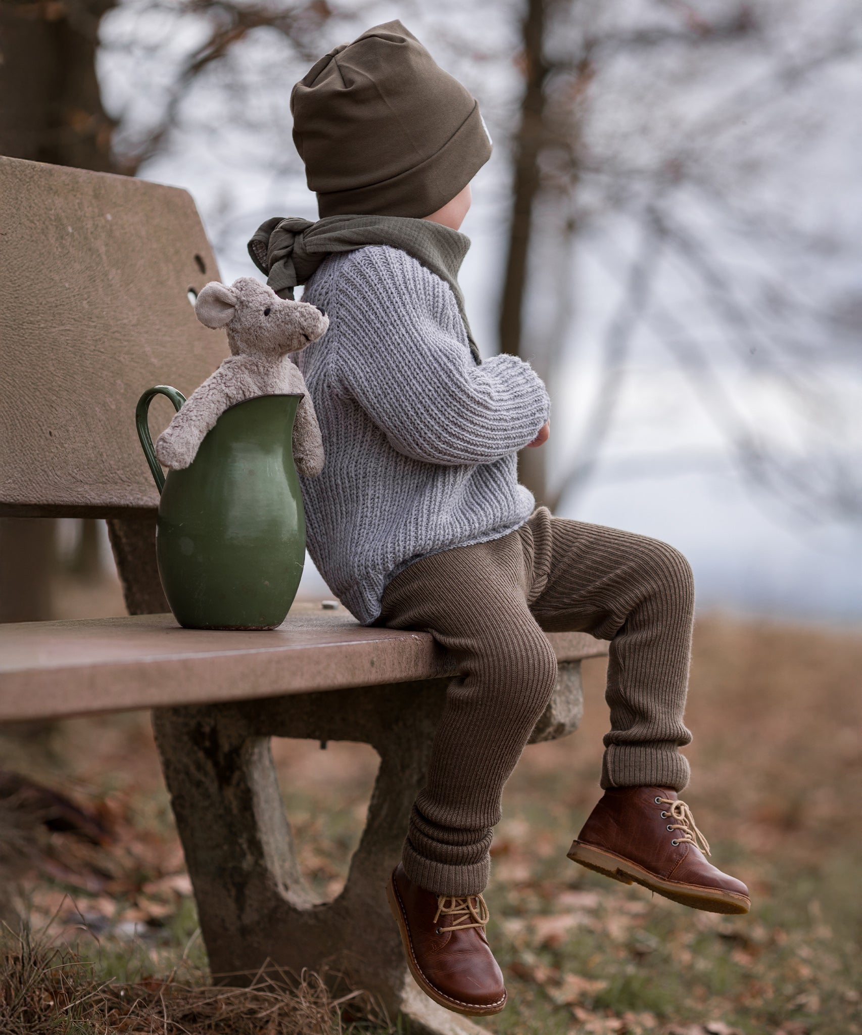 A child is sat on a light brown bench whilst looking off into the distance. The Senger Baby Mouse is sat inside a green ceramic jug, which shows the top half of the mouse. The jug is placed next to the child on the bench