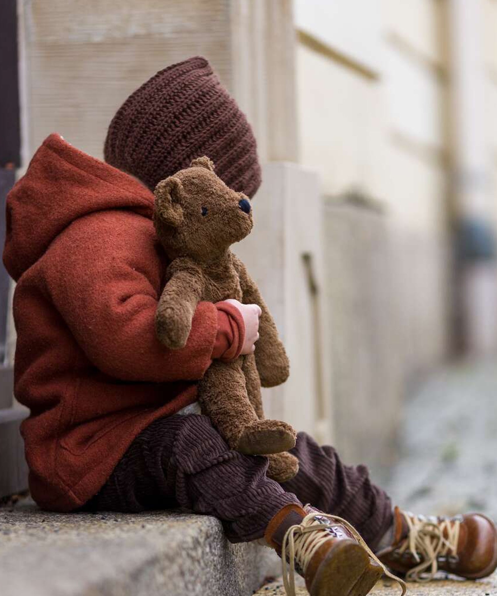 A child cuddling the Senger Small Floppy Brown Bear whilst sat on a step