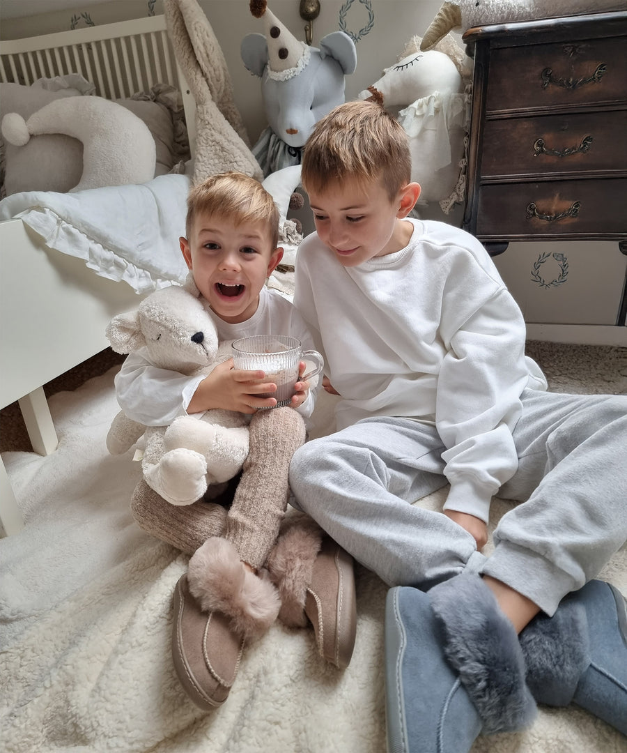 Two children sat on a white fluffy rug in their room. The child on the left is holding the Senger Large Cuddly Polarbear in their arms and a cup of hot chocolate