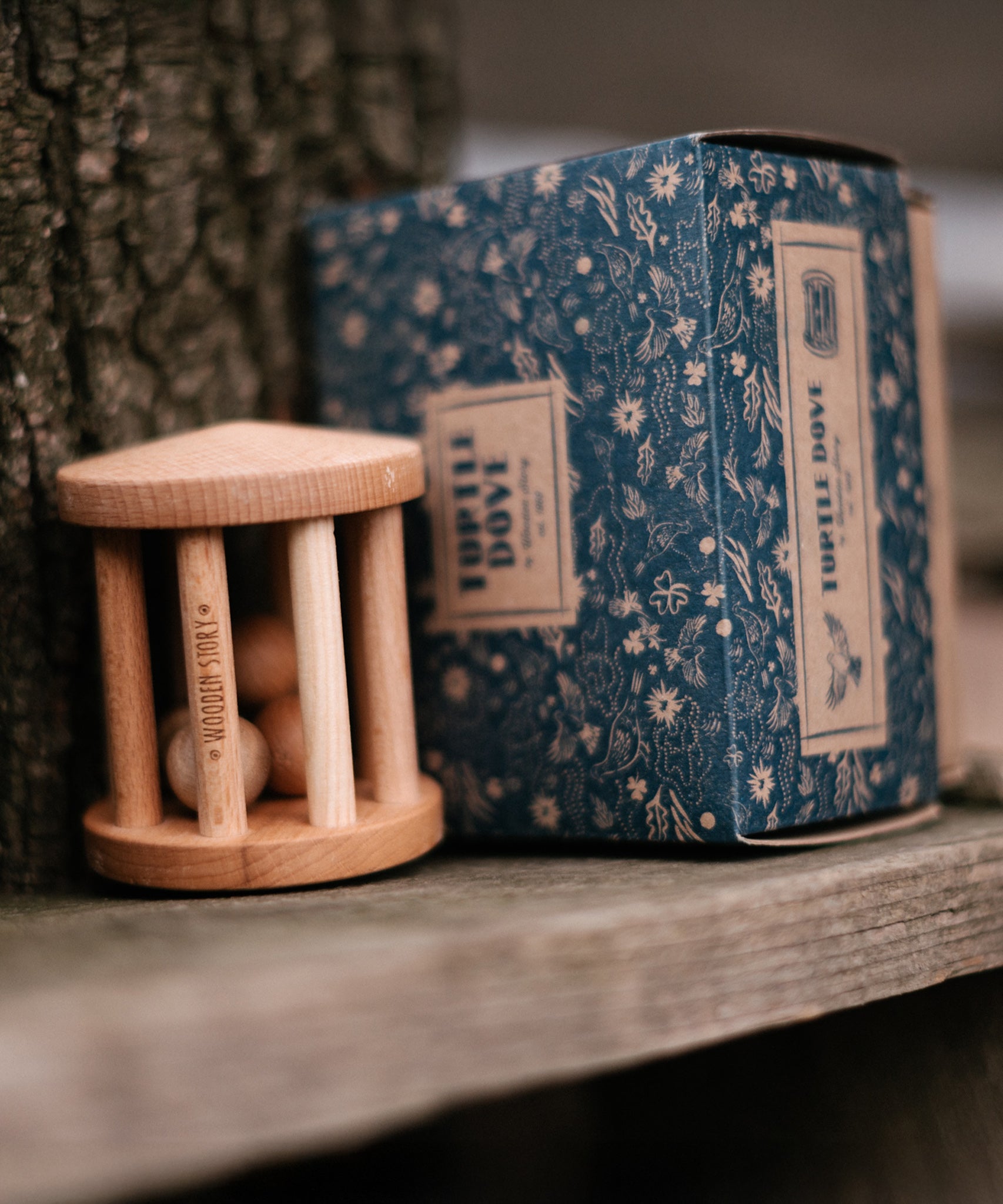 The Wooden Story Turtle Dove Rattle with its cardboard box next to is, is sat on a wooden plank next to a tree trunk