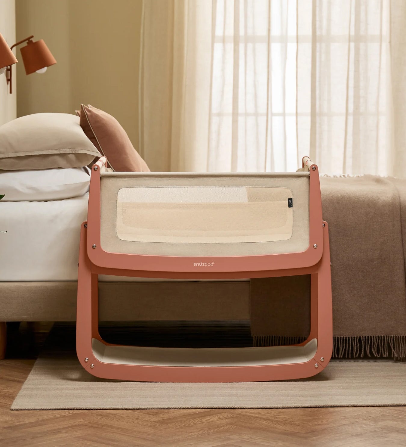 The SnuzPod 5 bedside baby crib with a lovely terracotta pink wooden frame, next to an adult's bed in a beautifully decorated bedroom.