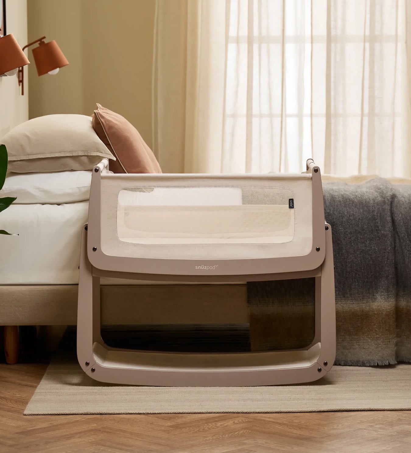 The SnuzPod 5 bedside baby crib with a lovely pale brown wooden frame, next to an adult's bed in a beautifully decorated bedroom.