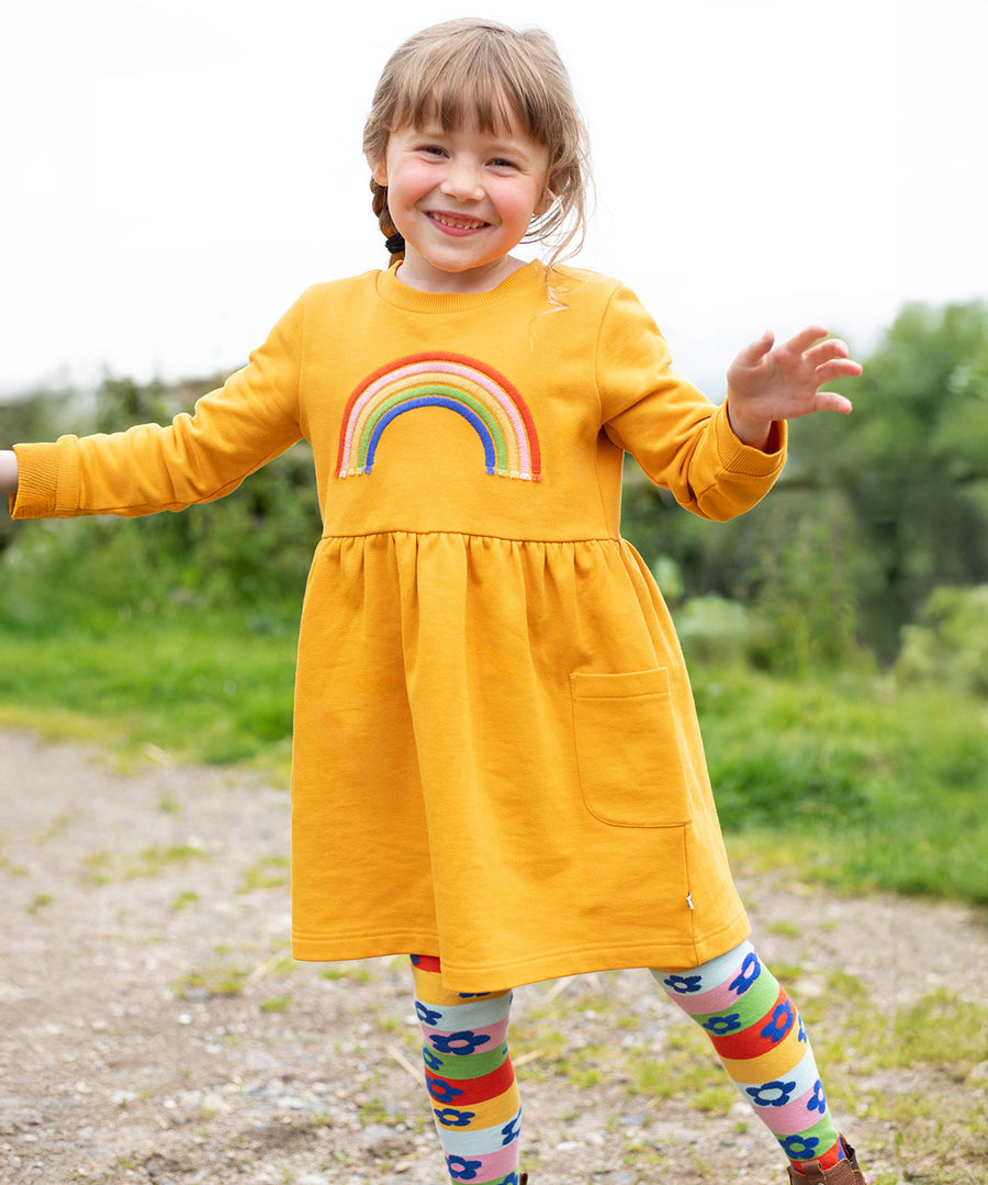 A child happily wearing the Frugi Niamh Dress - Gold/Rainbow. The dress is paired with the Frugi Retro Happy Norah Tights