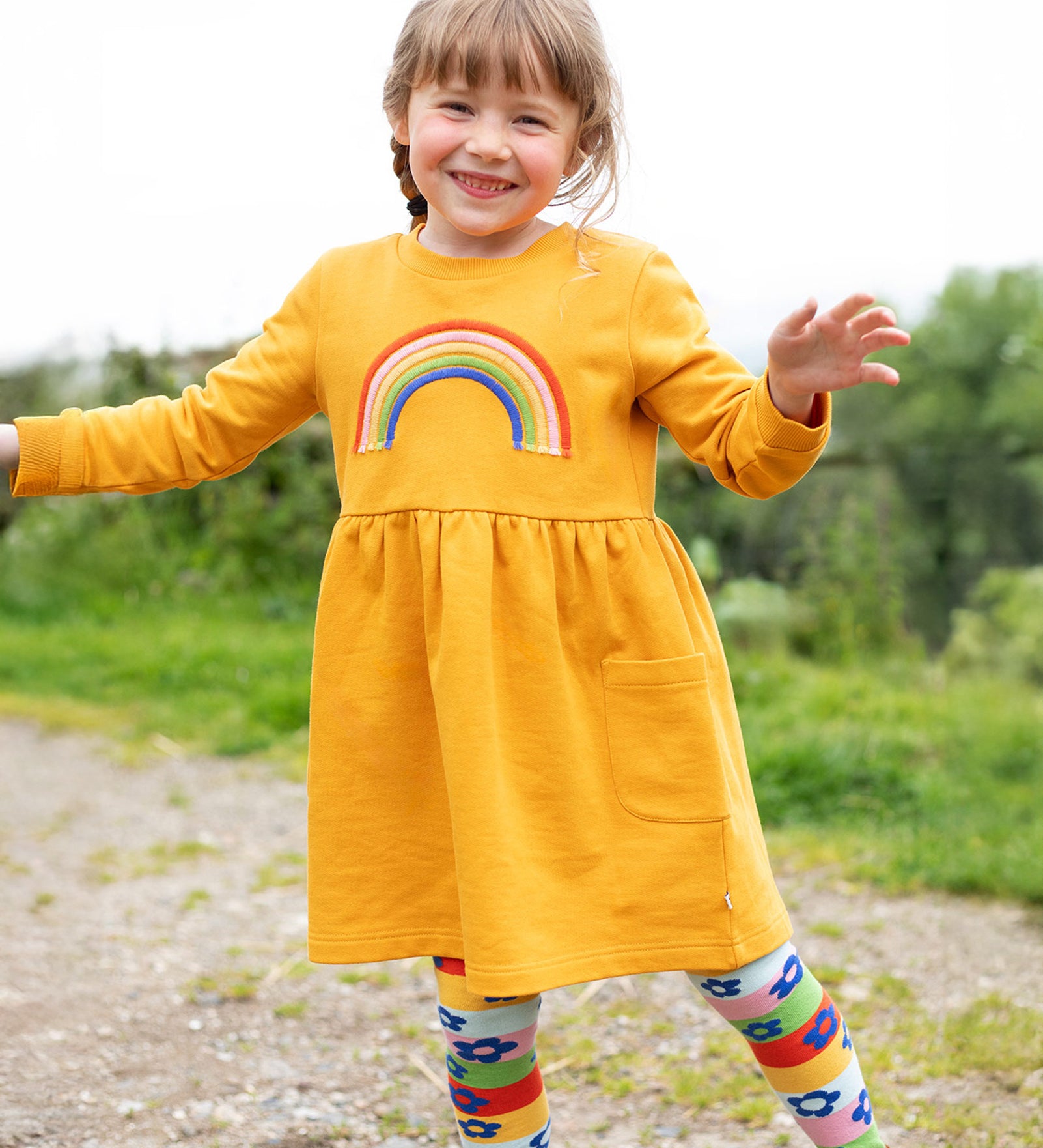 A child happily wearing the Frugi Niamh Dress - Gold/Rainbow. The dress is paired with the Frugi Retro Happy Norah Tights