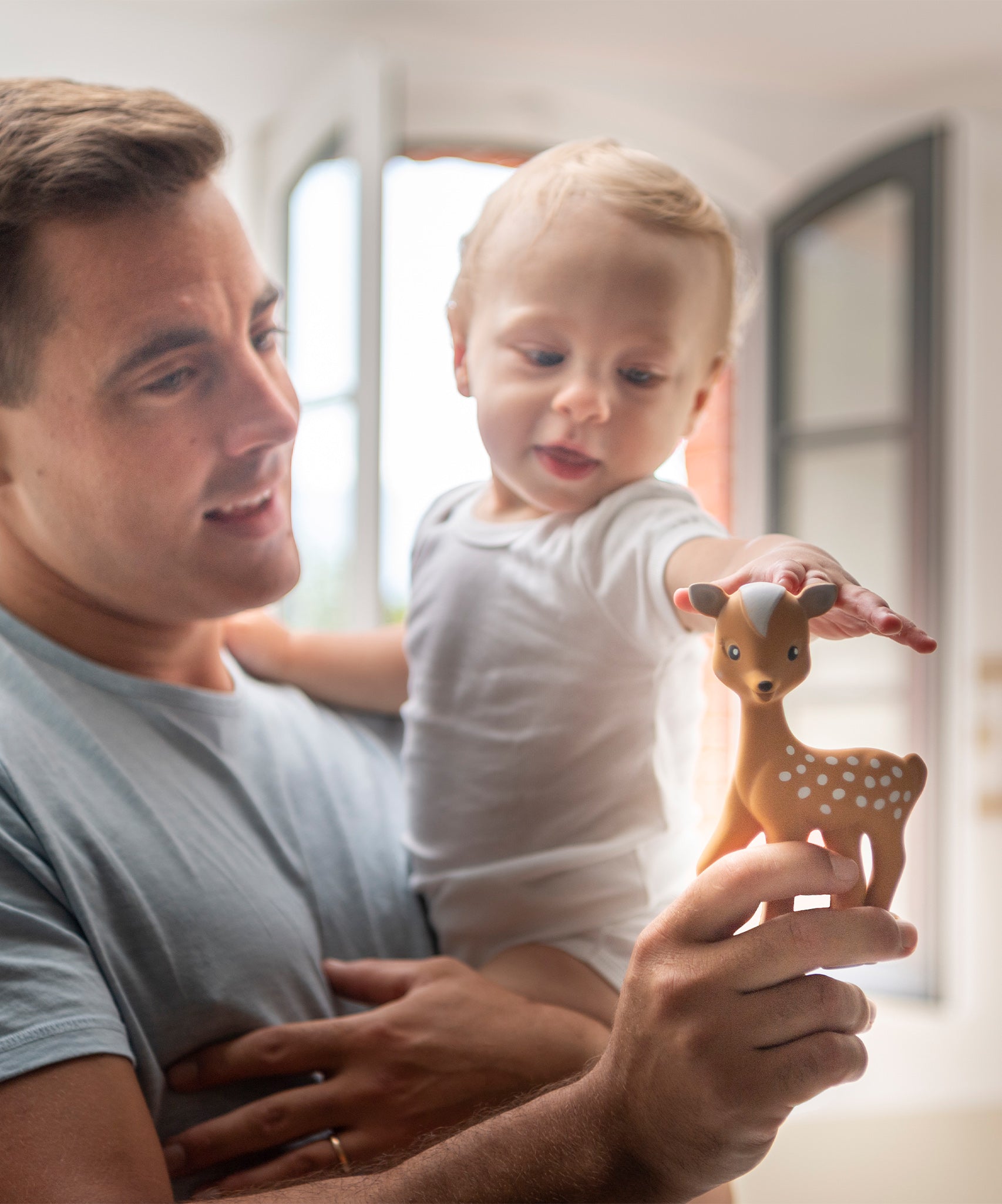 A child being held by an adult, whilst reaching out towards the Fanfan The Fawn Baby Teether, being held in the parents hand