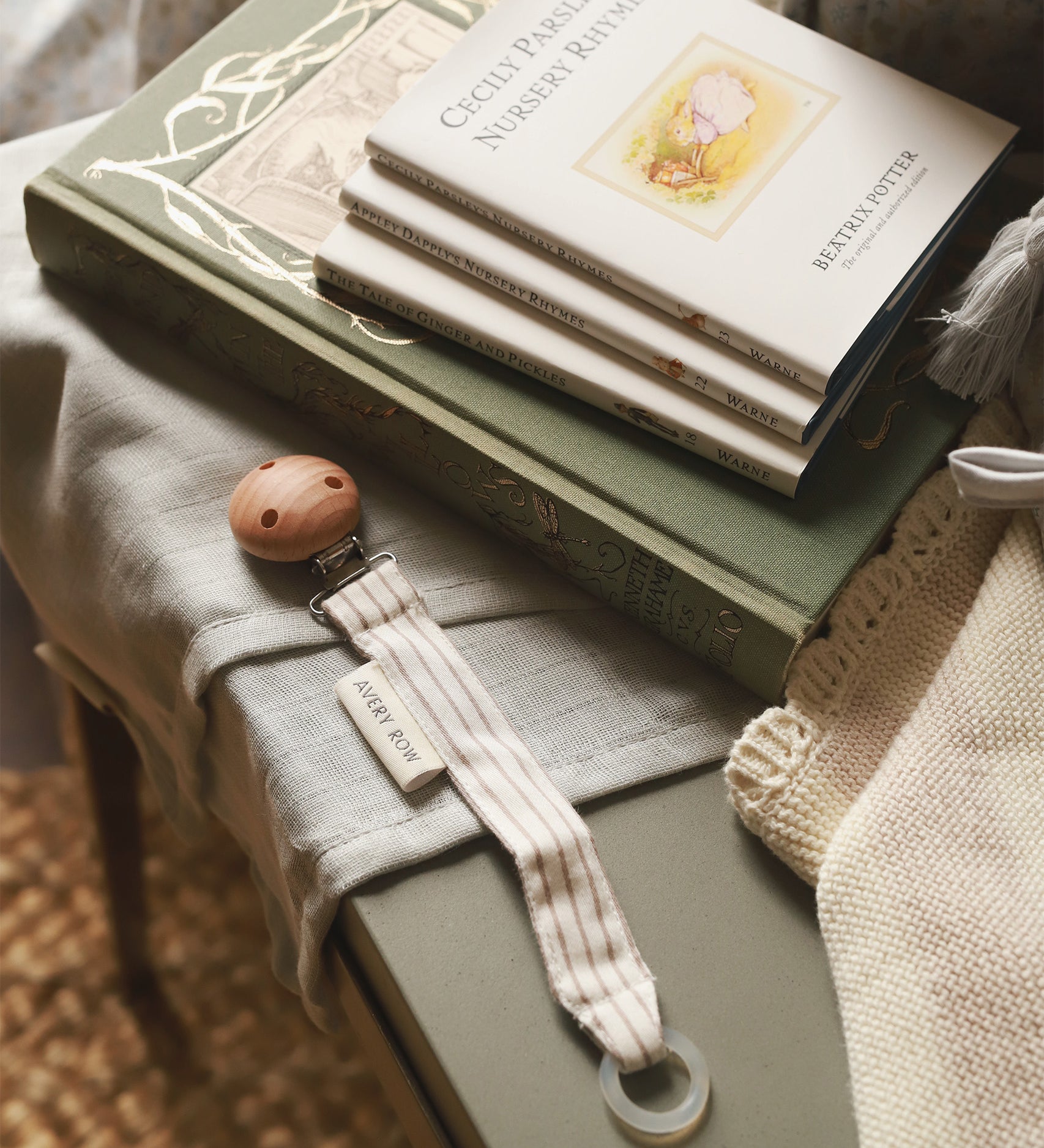 Avery Row Pacifier Holder in Stripe, next to a small pile of Beatrix Potter books and baby blankets