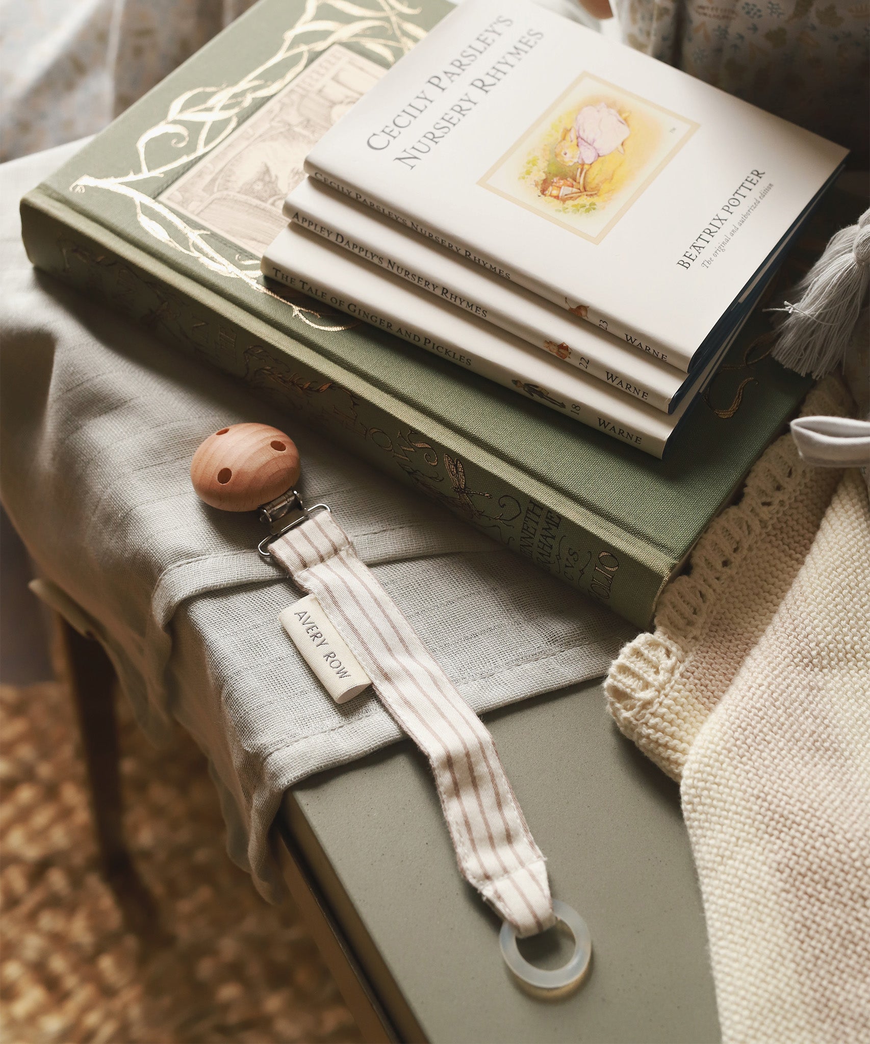 Avery Row Pacifier Holder in Stripe, next to a small pile of Beatrix Potter books and baby blankets