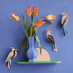 The Studio Roof Woodpecker, Jay and Hoopoe birds set on a blue coloured wall. The image also shows a blue vase filled with flowers on a small green shelf.