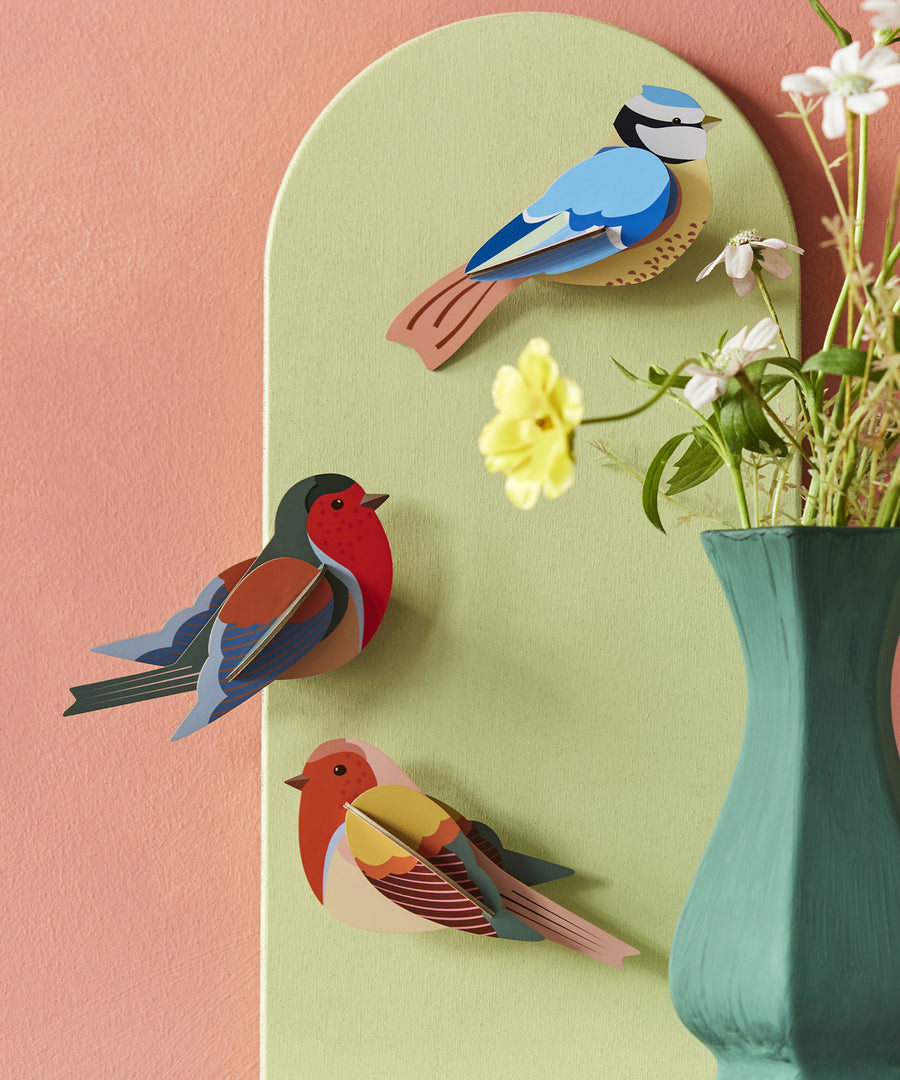 The Studio Roof Cardboard Robins and a cardboard Blue Tit on a pink and green background. The image also shows a green vase with flowers inside, to the right hand side of the image