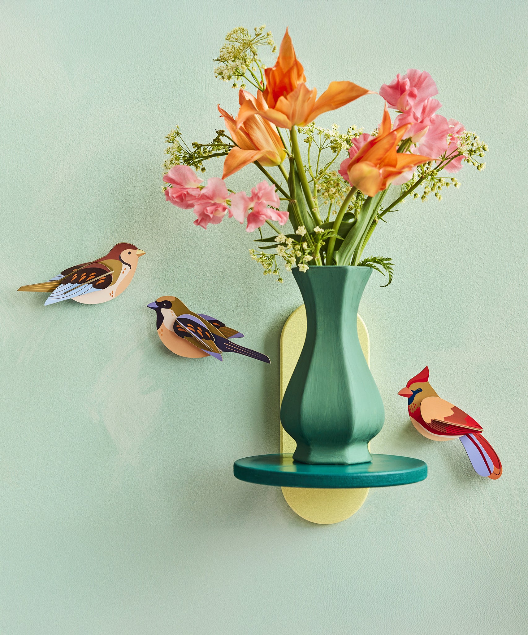 3 Studio Roof birds on a light green wall, with a vase full of colourful flowers on a small shelf also on a wall. The birds are the Sparrows and the Cardinal