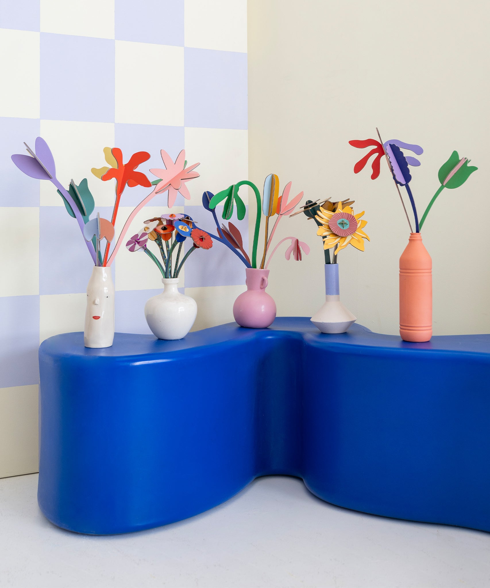 A collection of Studio Roof cardboard flowers in colourful vases, stood on a blue table