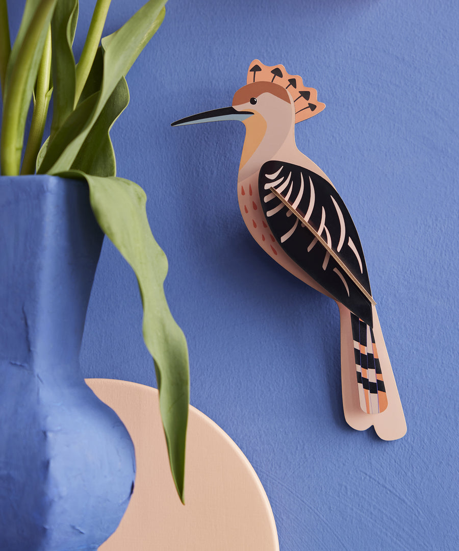 The Studio Roof Hoopoe Cardboard Model, set on a blue coloured background. On the left hand side of the image is a blue vase with a green plant inside.