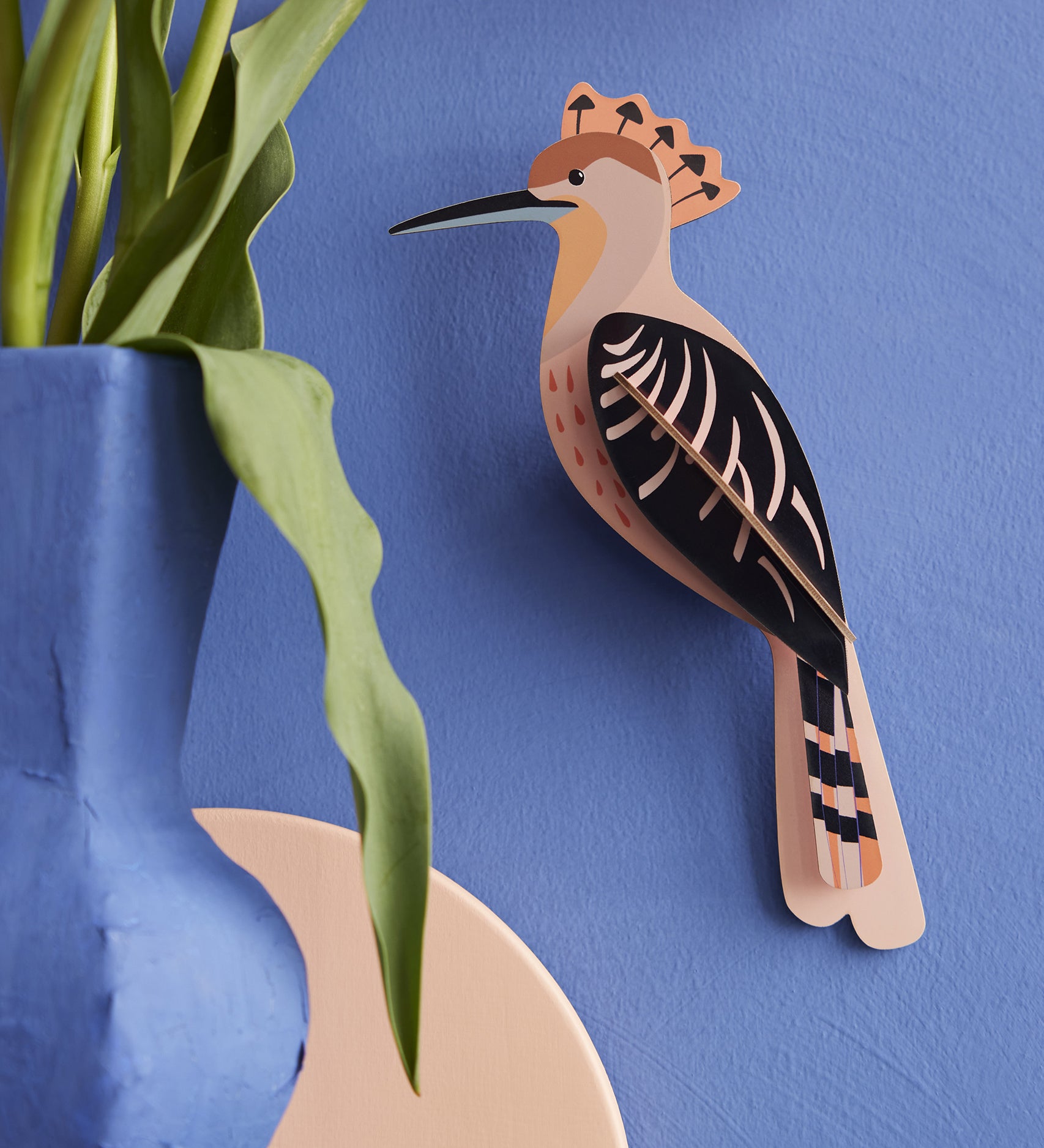 The Studio Roof Hoopoe Cardboard Model, set on a blue coloured background. On the left hand side of the image is a blue vase with a green plant inside.