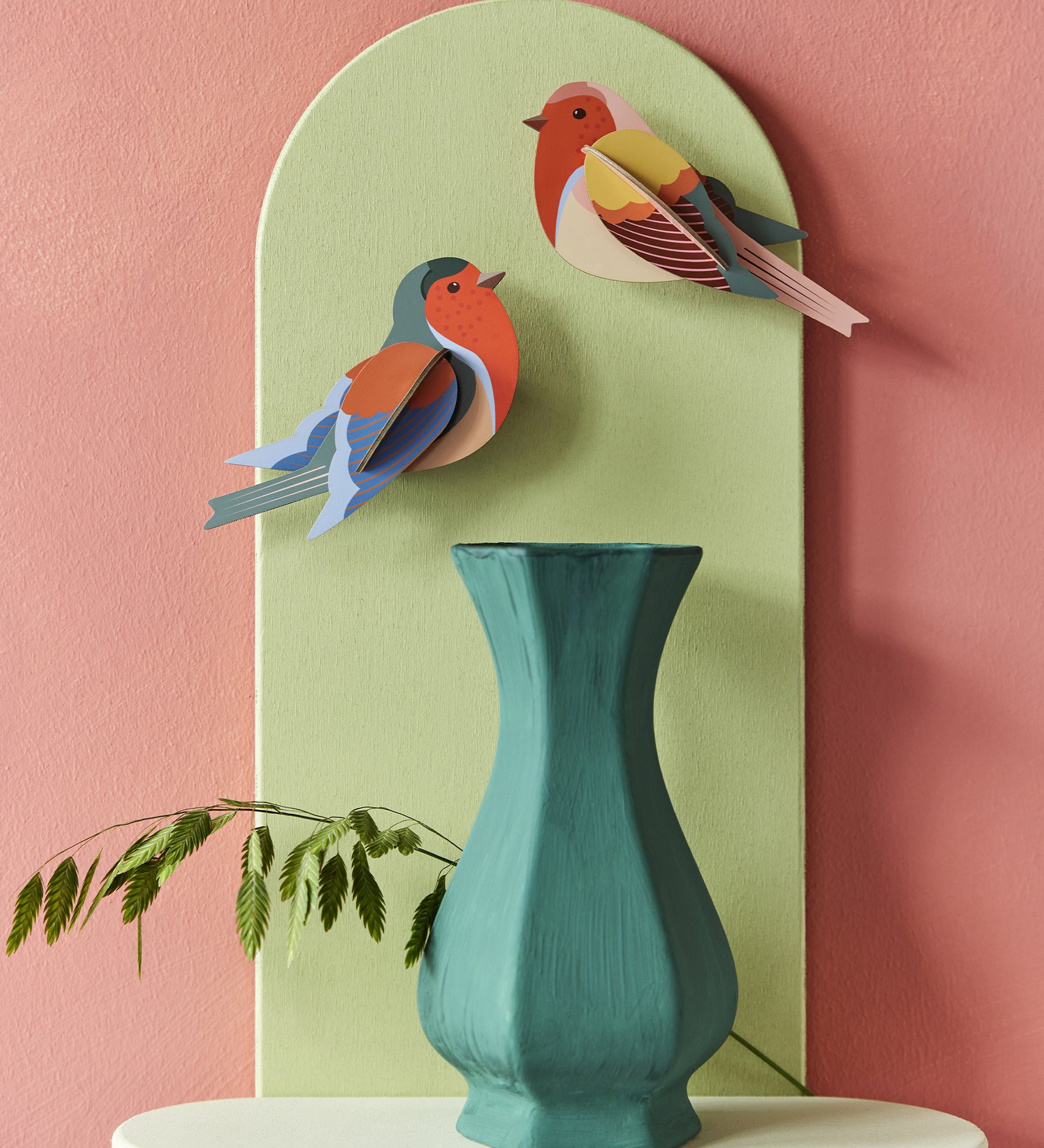 The Studio Roof Robins on a pink and green background, with a green vase and plant stem underneath the cardboard birds