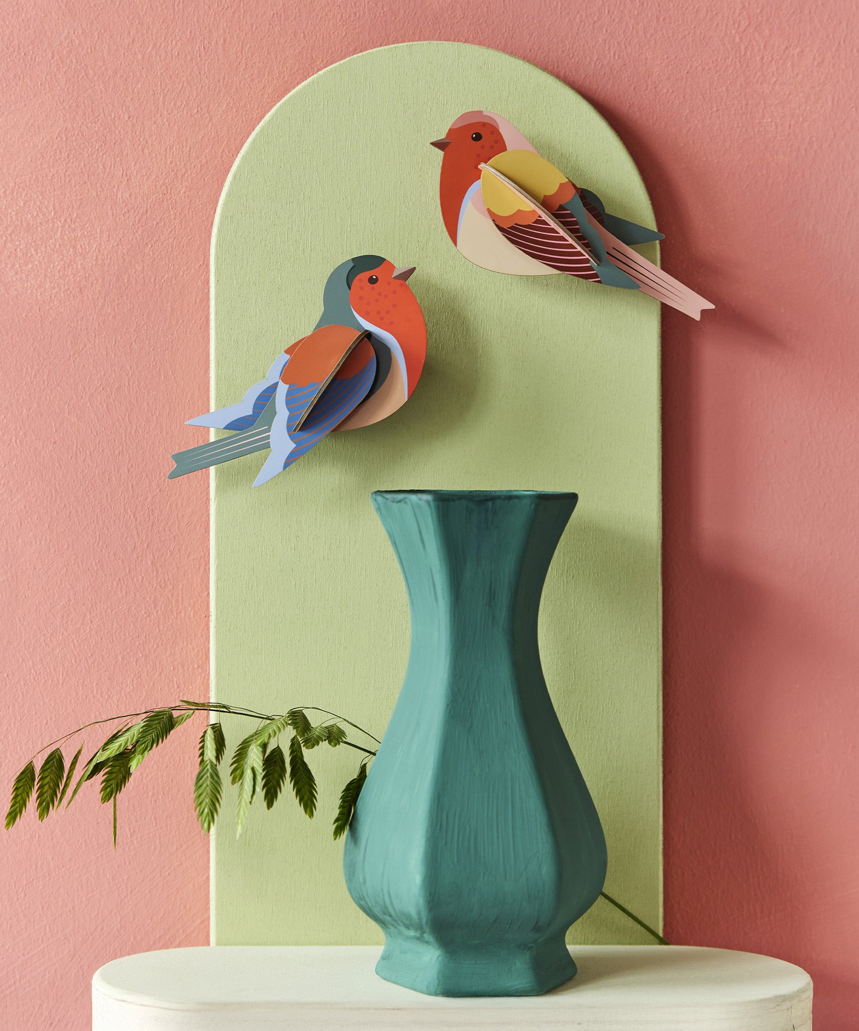 The Studio Roof Robins on a pink and green background, with a green vase and plant stem underneath the cardboard birds