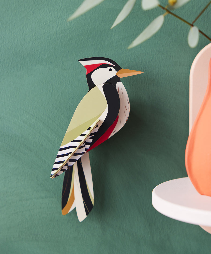 The Studio Roof Woodpecker Cardboard Model is set on a dark green background, with a peach ornament to the right hand side of the image