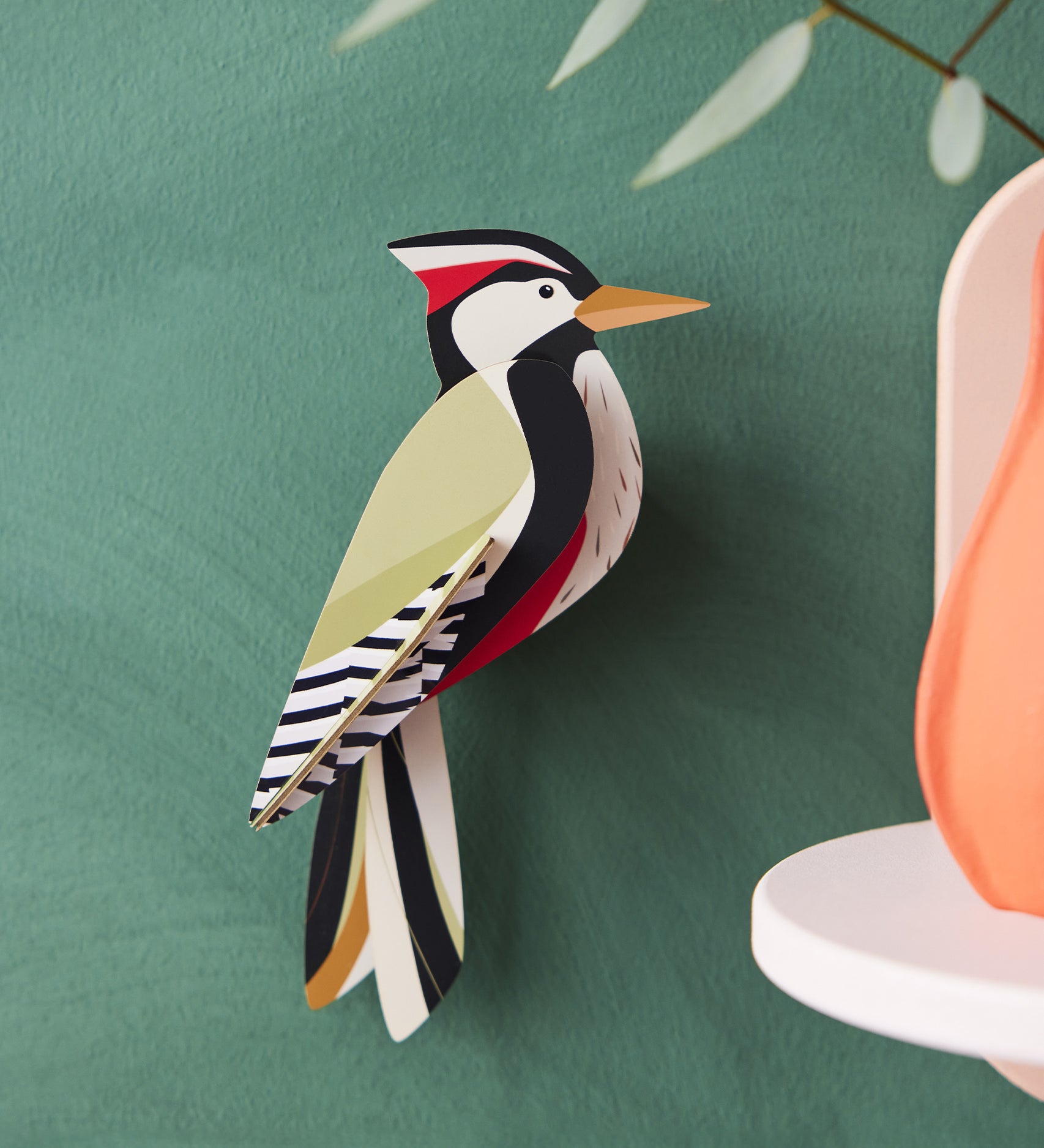 The Studio Roof Woodpecker Cardboard Model is set on a dark green background, with a peach ornament to the right hand side of the image