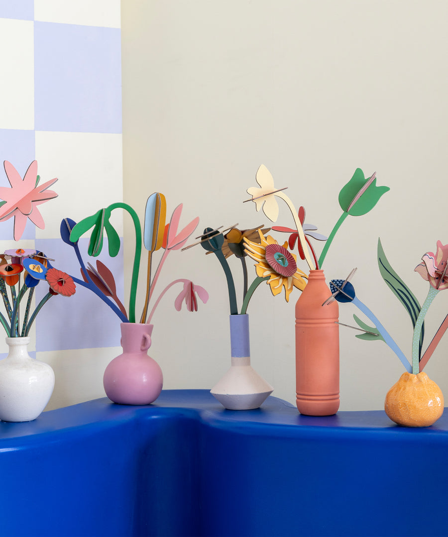 A collection of Studio Roof cardboard flowers in colourful vases, stood on a wooden table