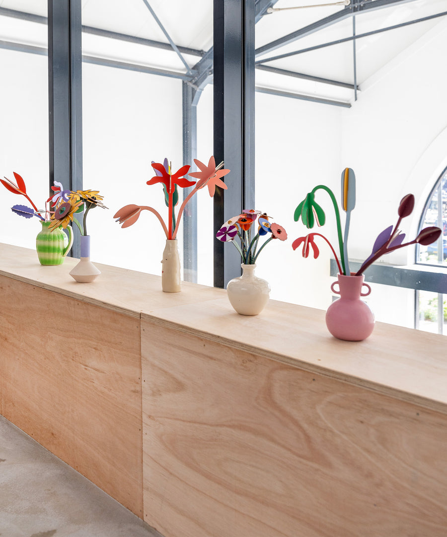 A collection of Studio Roof cardboard flowers in colourful vases, stood on a wooden table