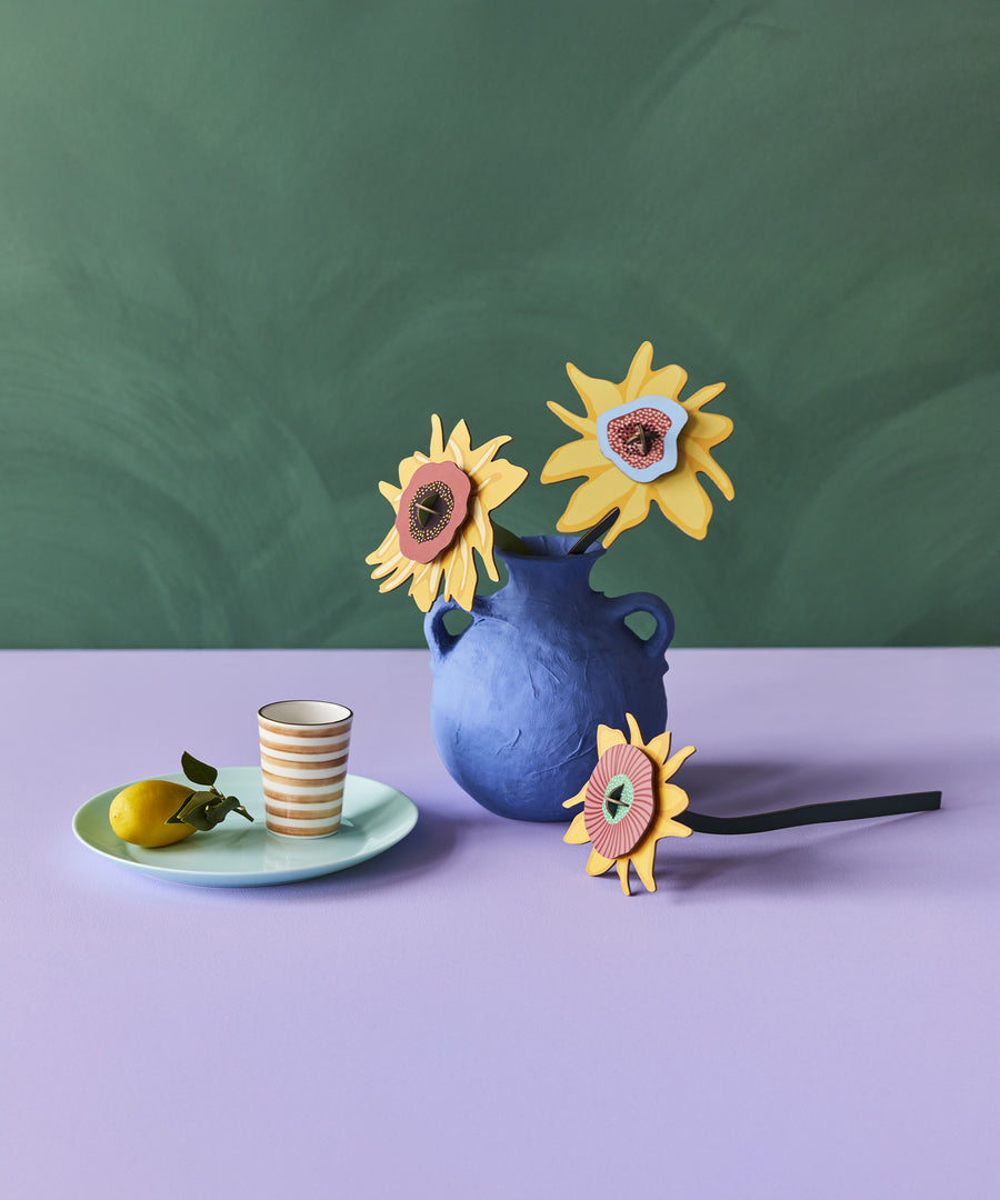 The Studio Roof Cardboard Bouquet - Van Gogh Sunflowers, stood in a blue vase. In front of the vase is one of the sunflwers laying horizontally and next to the vase on the left hand side is a small, light green plate with a lemon a a stripy cup. The image is on a blue and lilac background