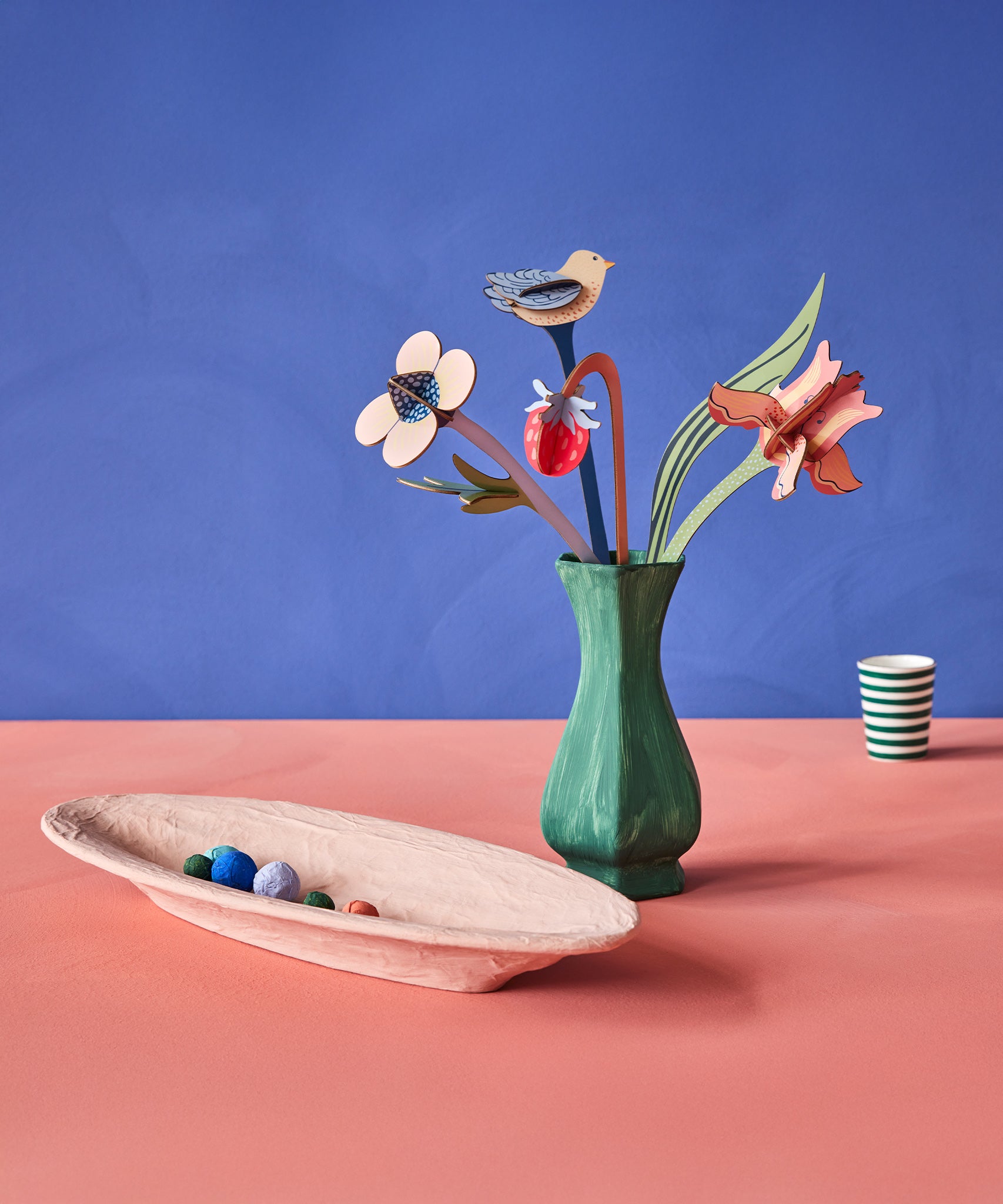 The Studio Roof Cardboard Bouquet - William Morris in a painted green vase, with a cream painted bowl with little papermache balls inside, and a green and white stripe cardboard cup in the background. The image is on a blue/purple and peach background