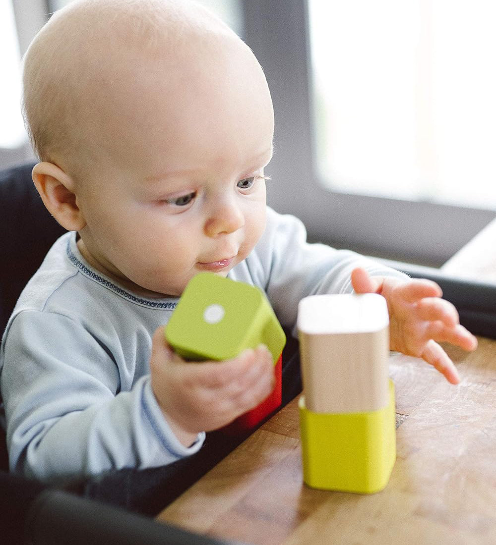 A baby playing with blocks from the  Tegu Baby's First Magnetic Blocks which is set on a wooden table. The baby is sat in a high chair at the table