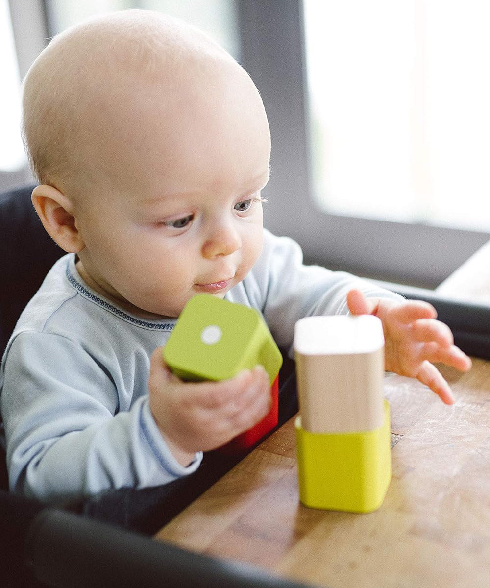 A baby playing with blocks from the  Tegu Baby's First Magnetic Blocks which is set on a wooden table. The baby is sat in a high chair at the table