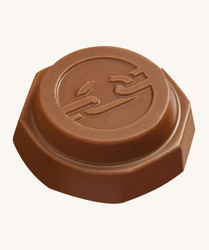 An angled view of the Tony's Chocolonely Tiny Mix chocolate button with the imprint of a broken chain on the front, to represent the breaking of slave labour. The image is on a cream background.