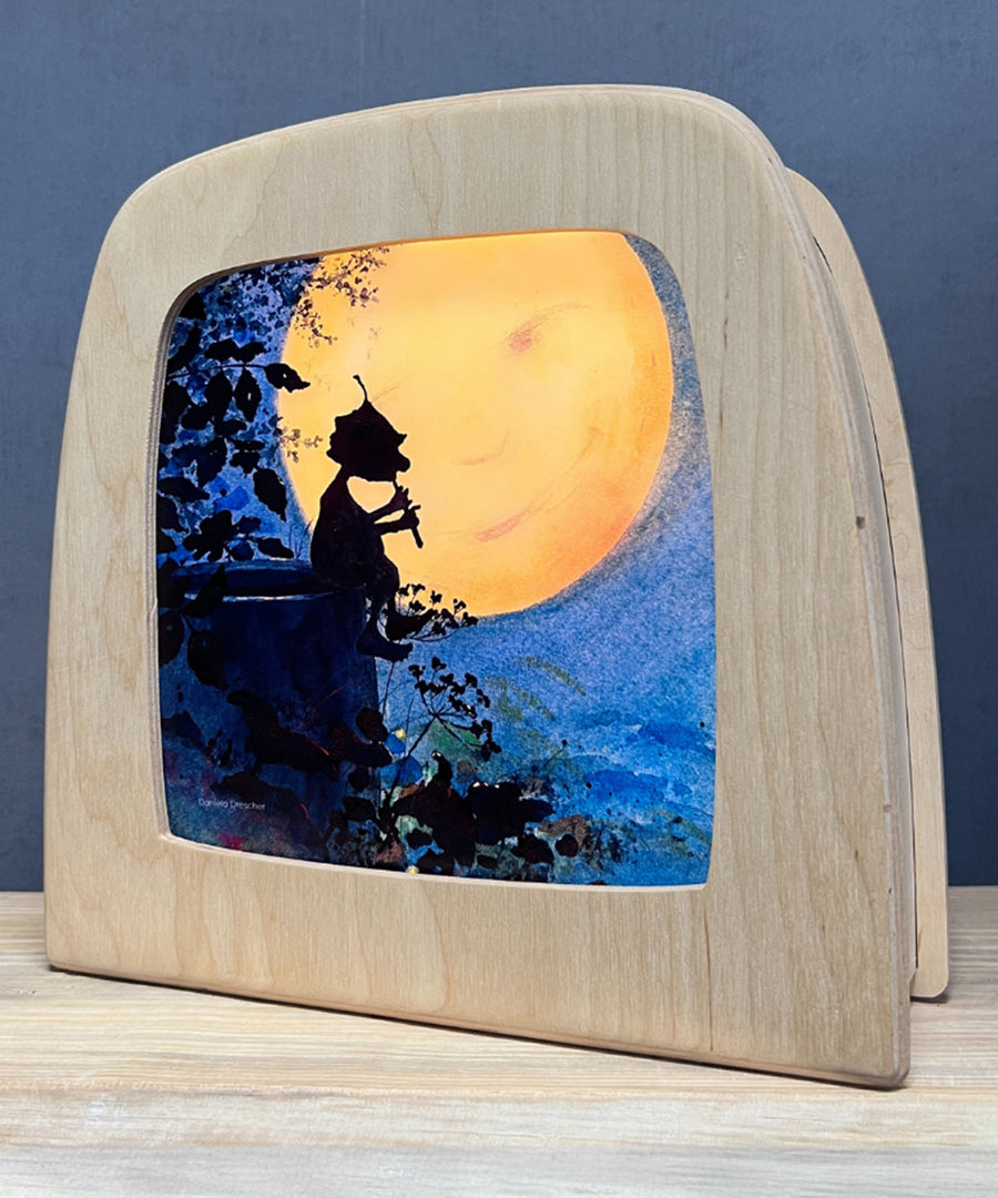 An illustration of a gnome playing a flute in the night with a full moon from the Adventures of Griswald Toverlux StoryLux Silhouette Sets, by Daniela Drescher. The illustration is inside a Magic Lamp which is stood on a table