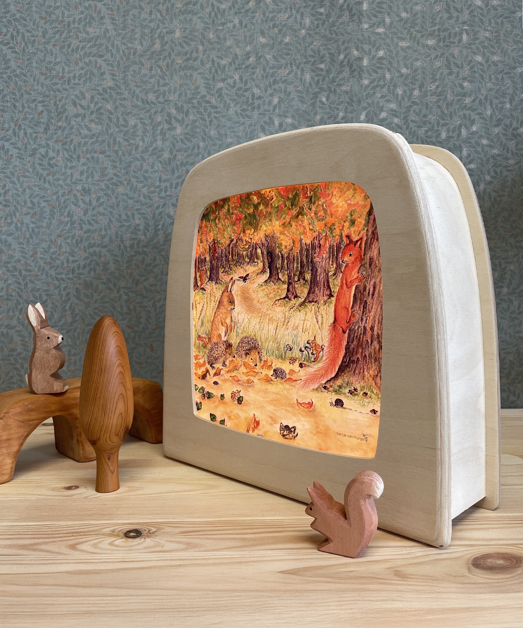 The Toverlux Magic Lamp with an illustration inside by Eentje Van Margo called Autumn Bliss. Surrounding the Magic Lamp are adorable wooden, woodland creatures. Everything is stood on a wooden dresser with patterned green wall paper in the background