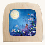 The Little Fairy Can't Sleep - Moon illustration by Daniela Drescher sat in a Toverlux Magic Lamp, on a cream background. The mage shows a fairy laying in bed underneath colourful flowers with a full moon in the sky