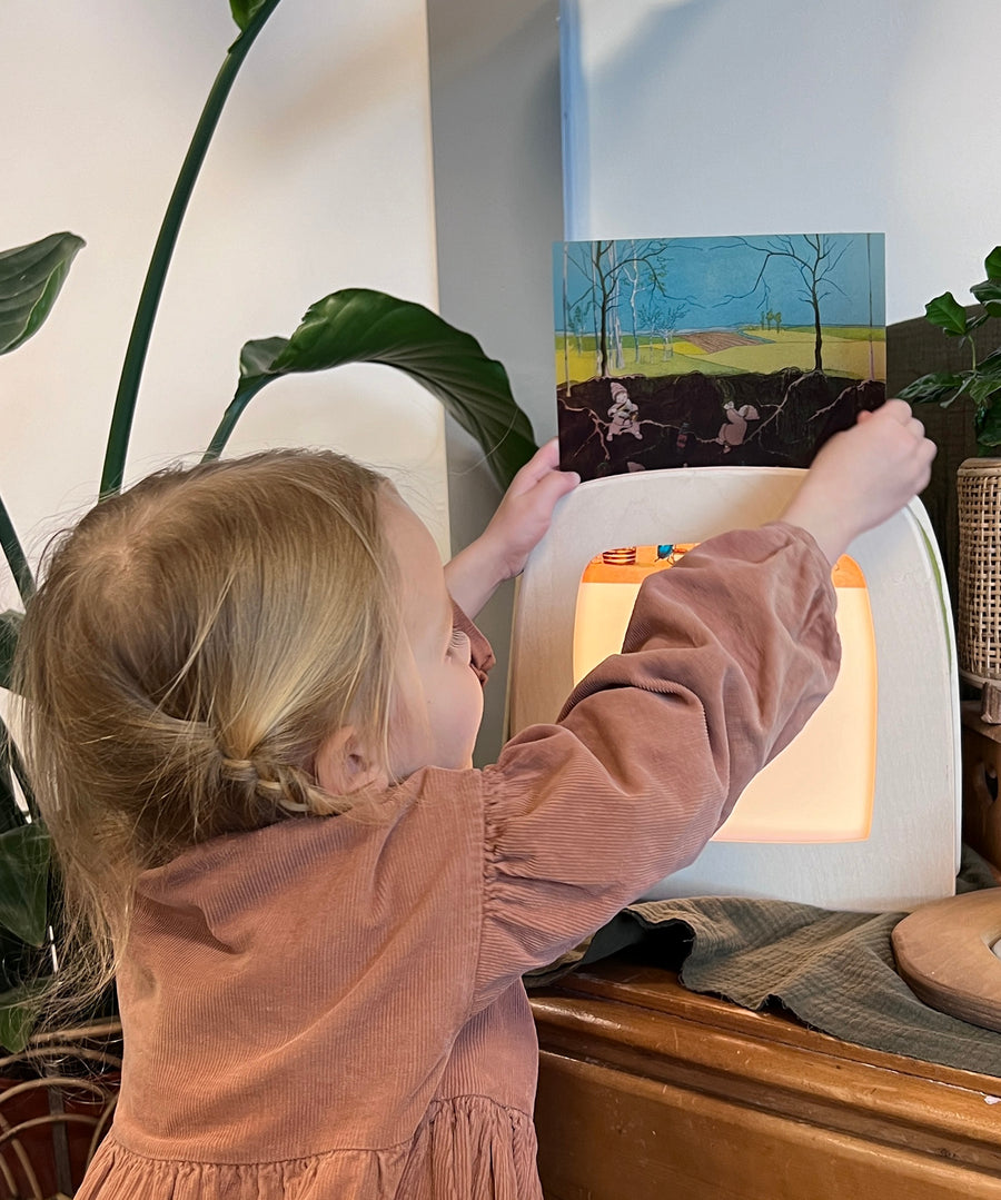 A child putting an illustration into the Toverlux Magic lamp, which is stood on a wooden dresser and is surrounded by green plants.