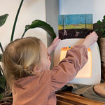 A child putting an illustration into the Toverlux Magic lamp, which is stood on a wooden dresser and is surrounded by green plants.