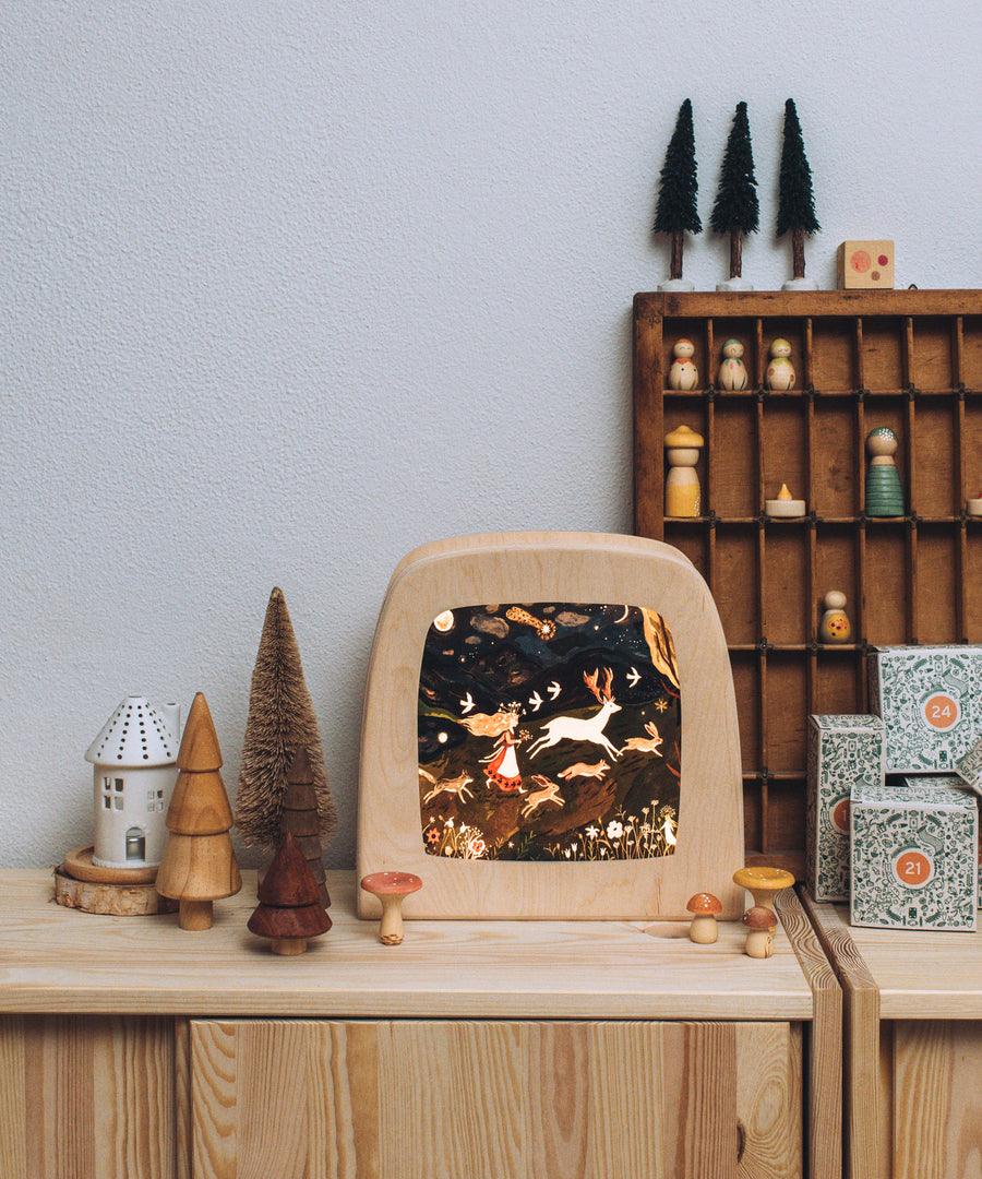 The Toverlux Magic Lamp with an illustration inside by Tijana Draws called Return Of Happiness. Surrounding the Magic Lamp are different wooden toys with everything stood on a light brown, wooden dresser