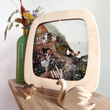 The Toverlux Artisan Magic Window Frame on the Frame base, with an illustration inside. The illustrations is by Robin Pieterse, called Rose Cottage. In front of the frame is a small wooden wan and bird toy, and behind the frame is a green vase with flowers inside