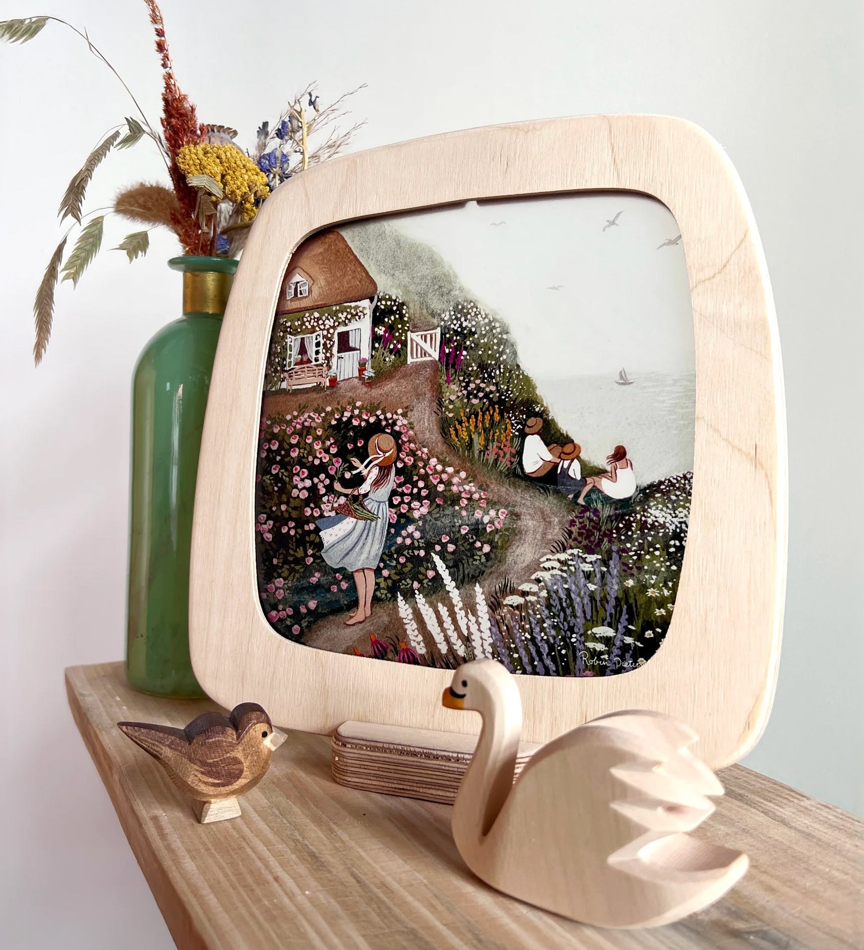 The Toverlux Artisan Magic Window Frame on the Frame base, with an illustration inside. The illustrations is by Robin Pieterse, called Rose Cottage. In front of the frame is a small wooden wan and bird toy, and behind the frame is a green vase with flowers inside