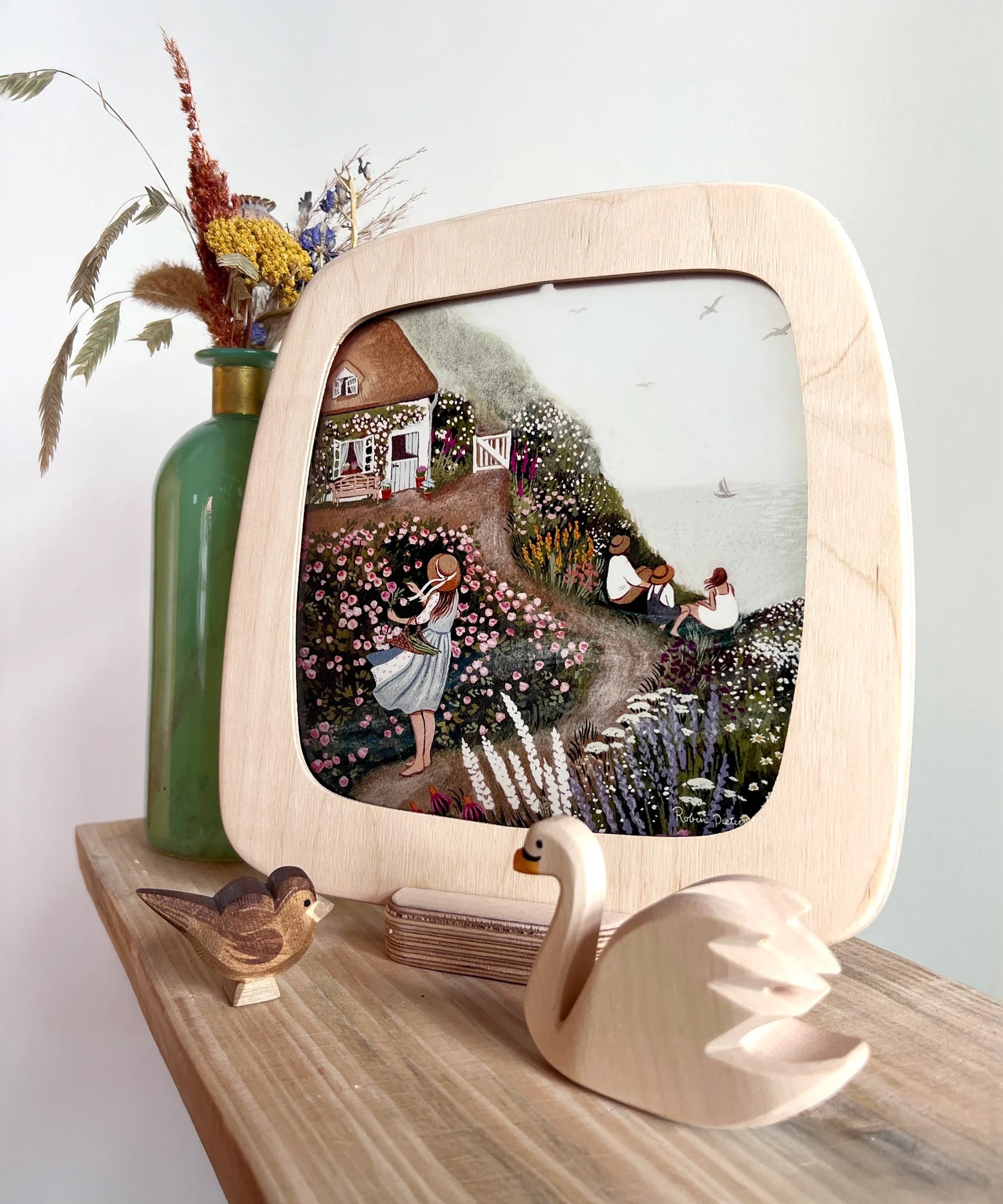 The Toverlux Artisan Magic Window Frame on the Frame base, with an illustration inside. The illustrations is by Robin Pieterse, called Rose Cottage. In front of the frame is a small wooden wan and bird toy, and behind the frame is a green vase with flowers inside