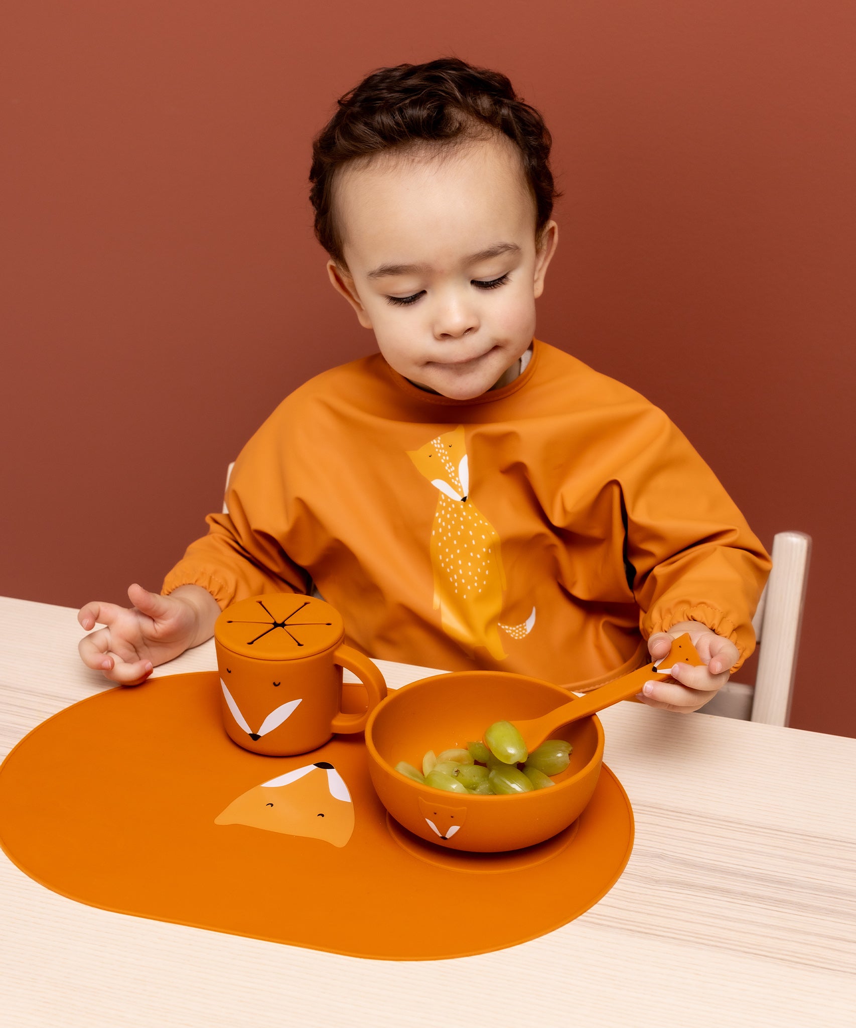 Child wearing the Trixie Animal Friends Waterproof Long Sleeve Bib, orange, Mr Fox, with matching tableware. 