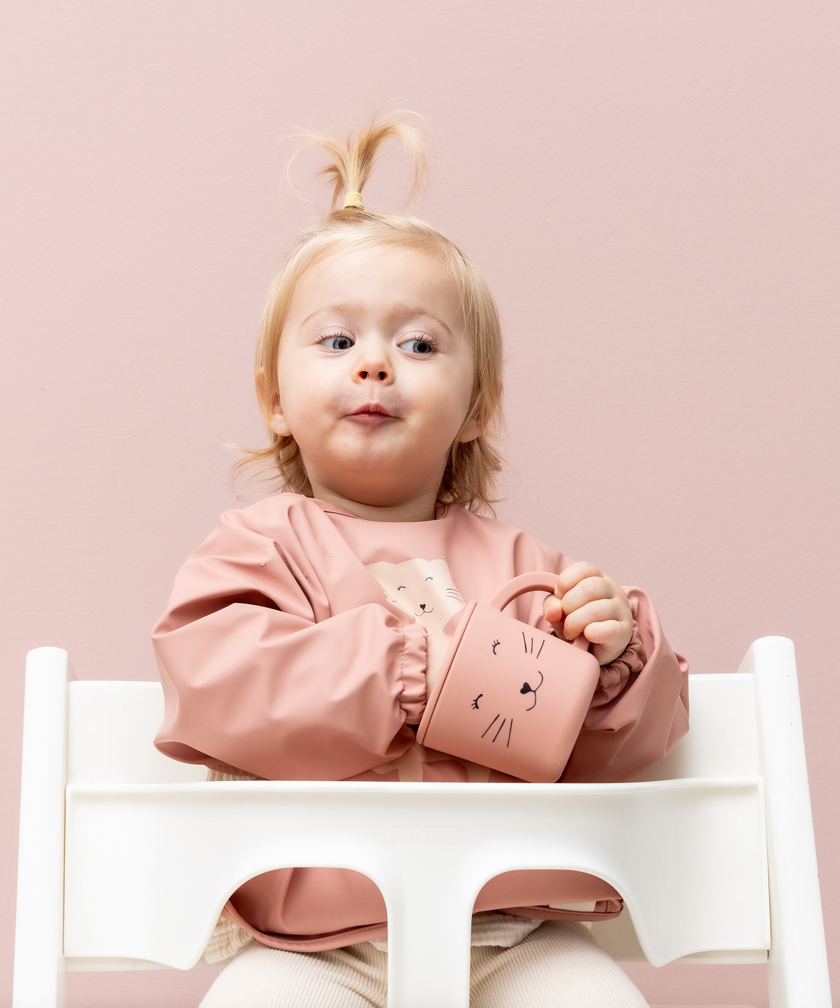 Baby sat in a high chair in a Trixie Animal Friends Waterproof Long Sleeve Bib, pink, Mrs Cat and matching sippy cup. 