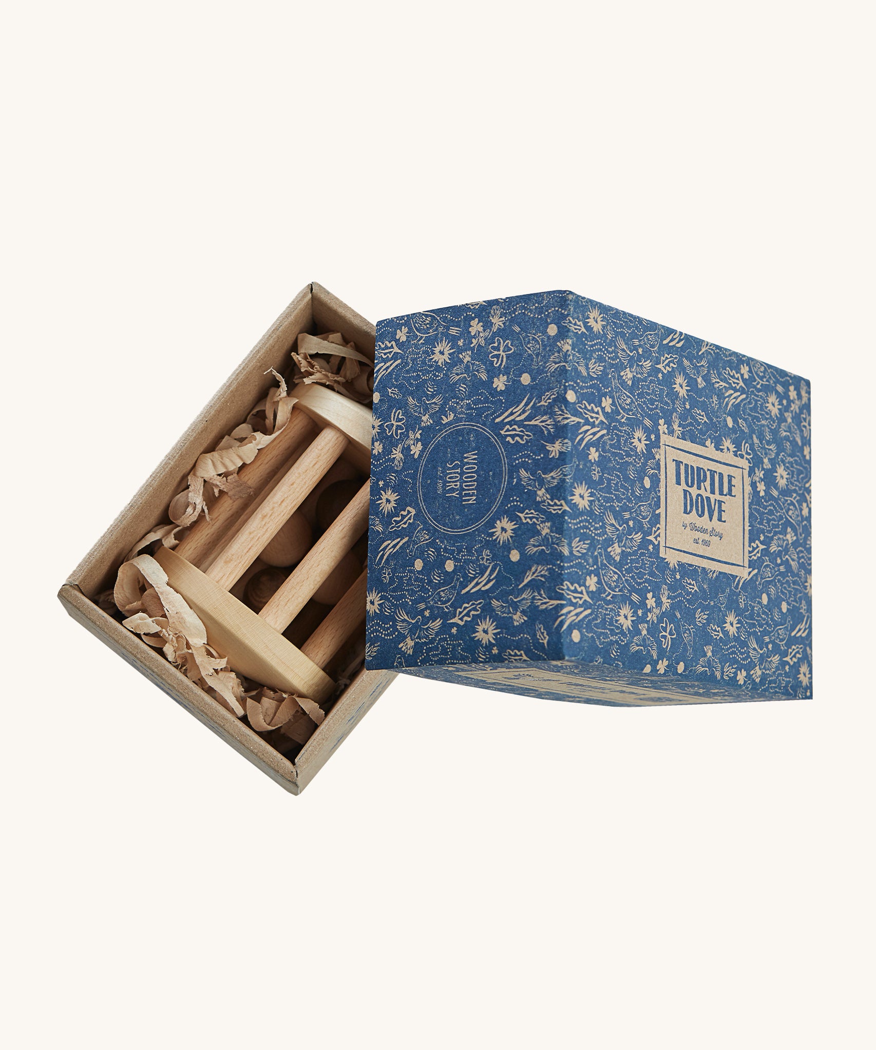 The Wooden Story Turtle Dove Rattle inside its cardboard box packaging. The rattle is laid on wood shavings